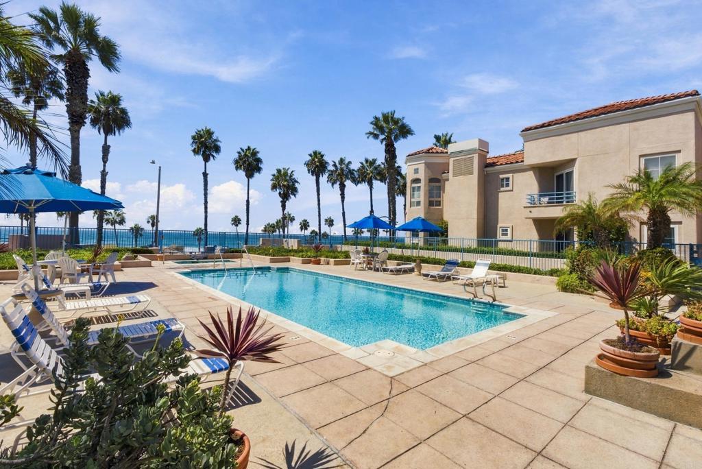 Outdoor pool with lounge chairs and palm trees, overlooking the ocean beside a coastal condominium.