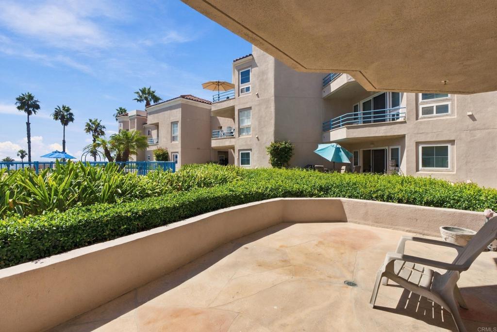 Condominium patio with lounge chairs, a turquoise umbrella, clipped hedges, and palm trees.