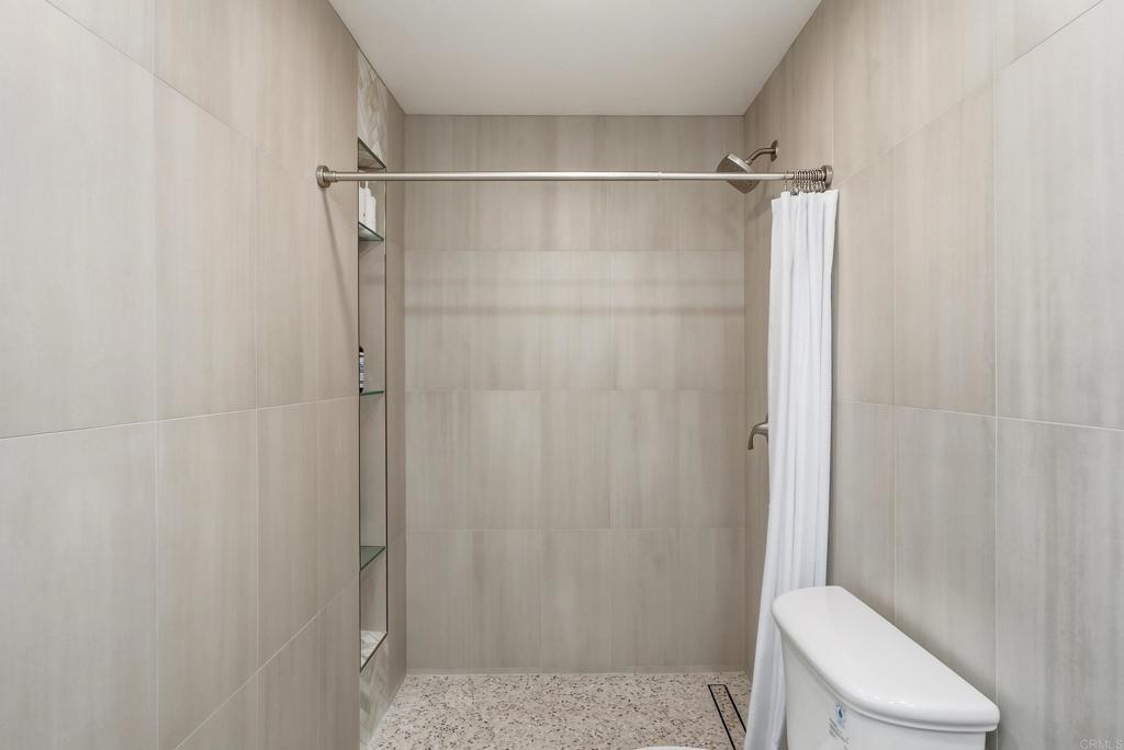 Walk-in shower with pale tile walls, pebble-pattern flooring, glass shelves, and a white curtain.