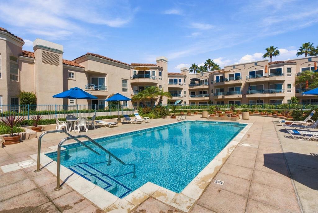 Condominium pool courtyard with blue umbrellas, lounge chairs, and surrounding apartment buildings.