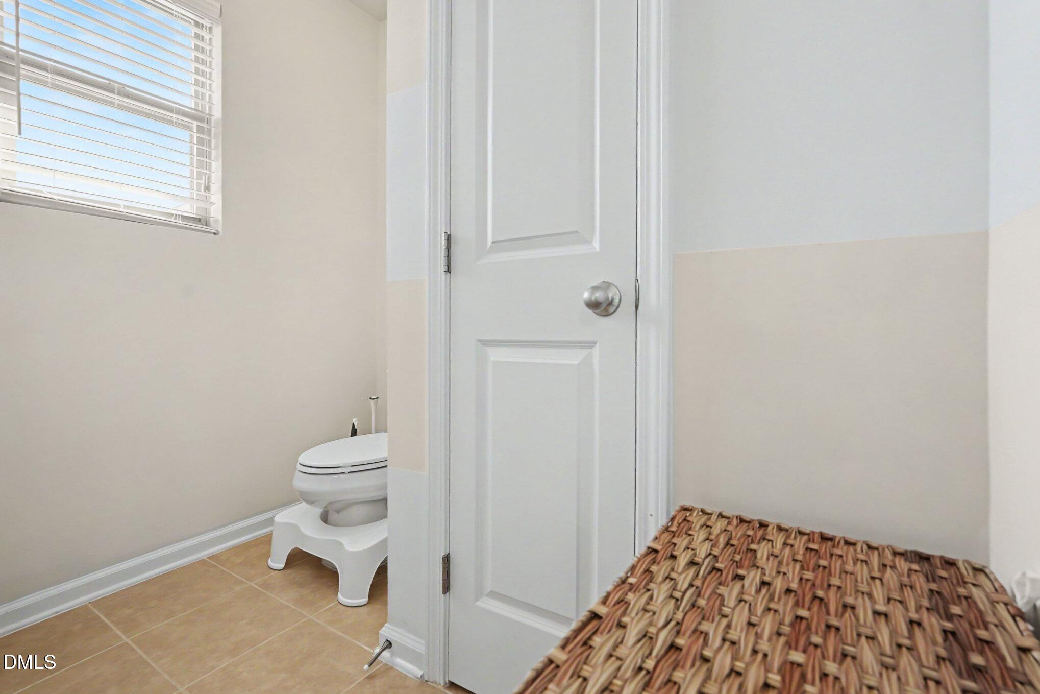 Bathroom with a toilet beneath a window, beige tile floor, and white paneled door.
