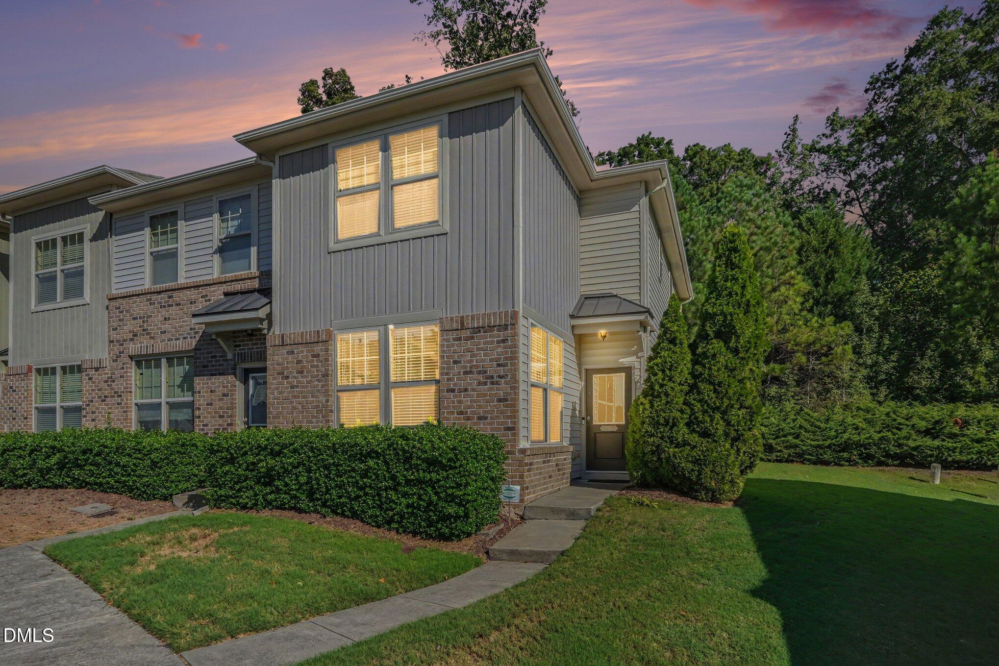 Two-story brick and gray-sided home with lit windows, trimmed hedges, and a curved front walk at dusk.