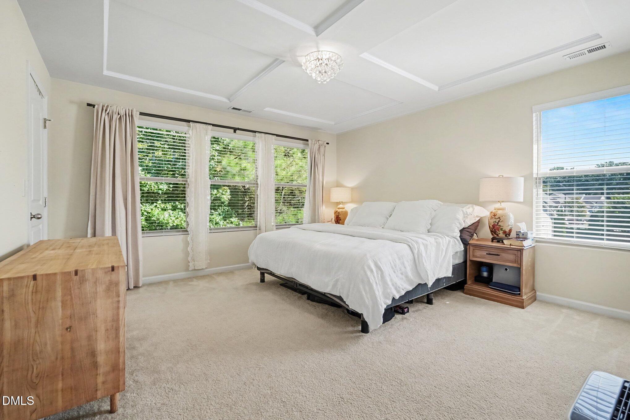 Bedroom with a white bed, tray ceiling, crystal chandelier, and windows overlooking greenery.