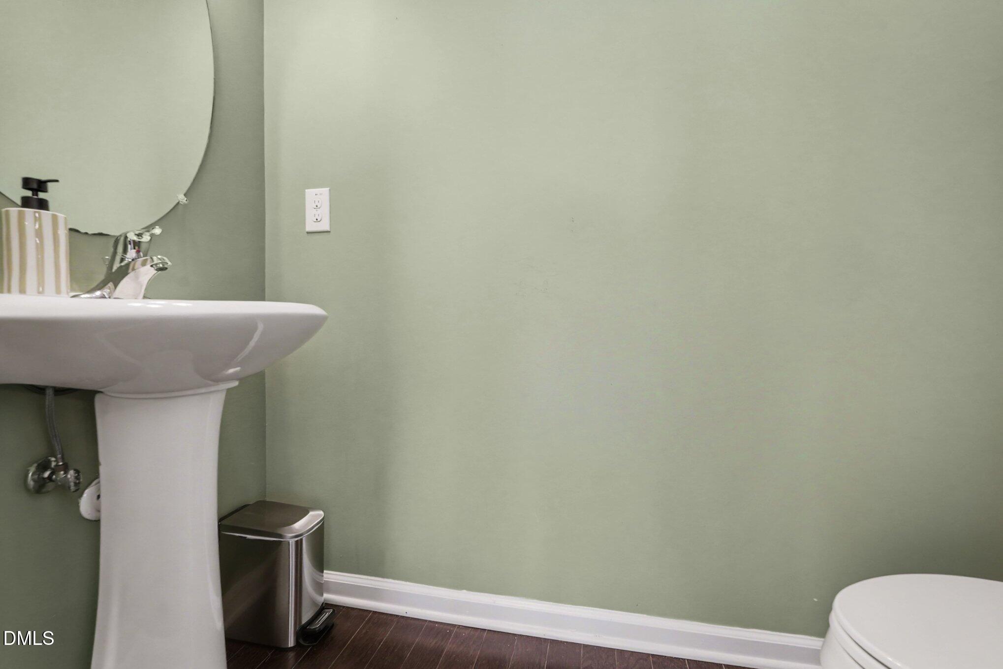 Bathroom with a white pedestal sink, round mirror, pale green walls, and dark wood flooring.