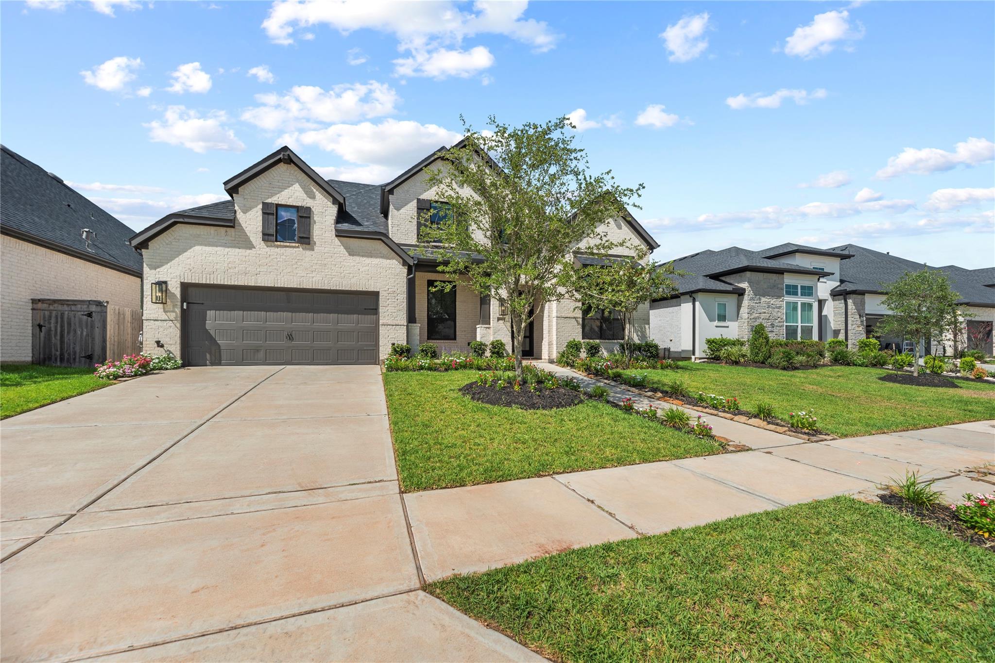Two-story light brick house with a dark double garage, landscaped front yard, and wide concrete driveway.