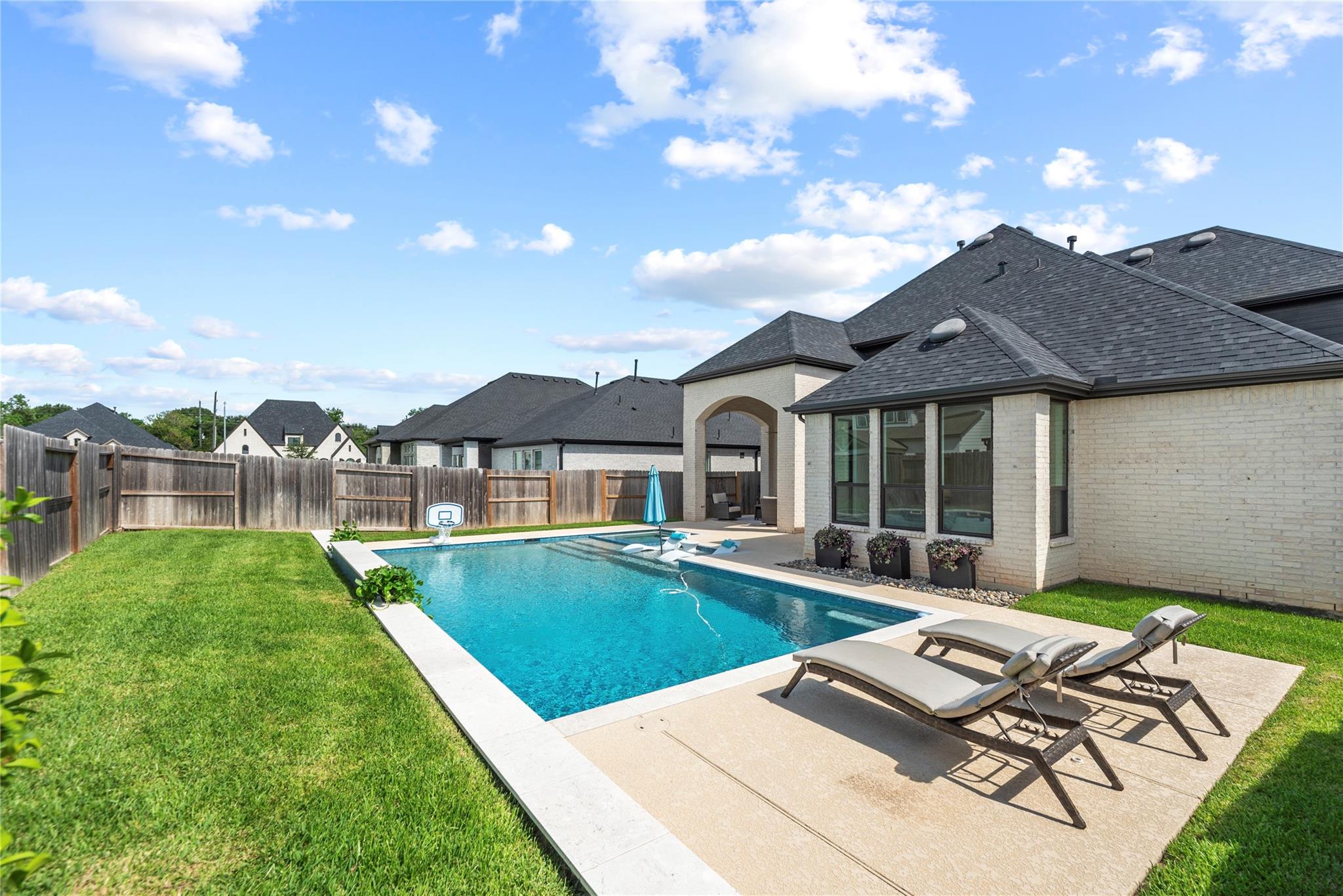 Backyard pool with basketball hoop, floating loungers, and a paved patio beside the brick house.