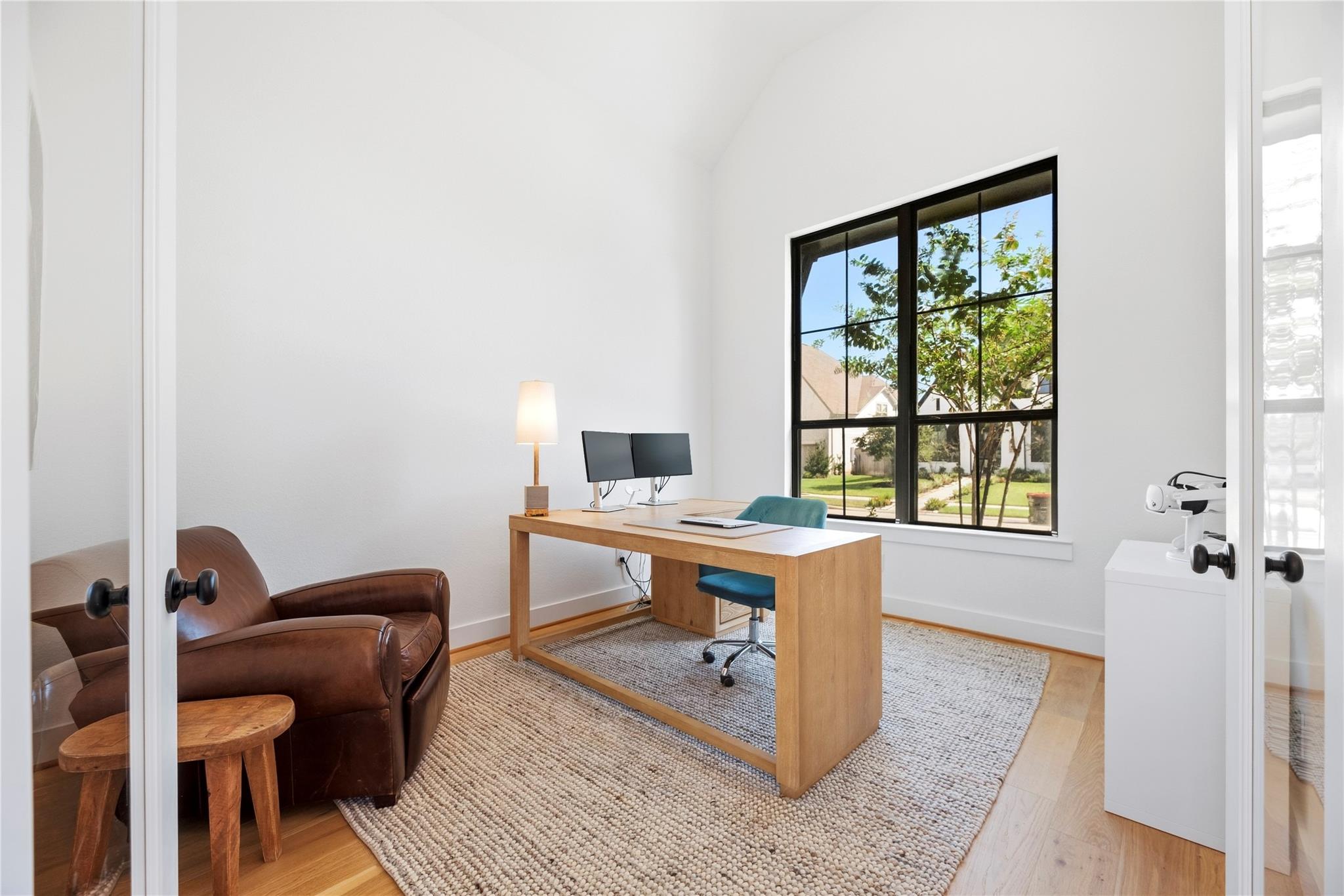 Bright home office with a wood desk and dual monitors beside a large black-framed window.