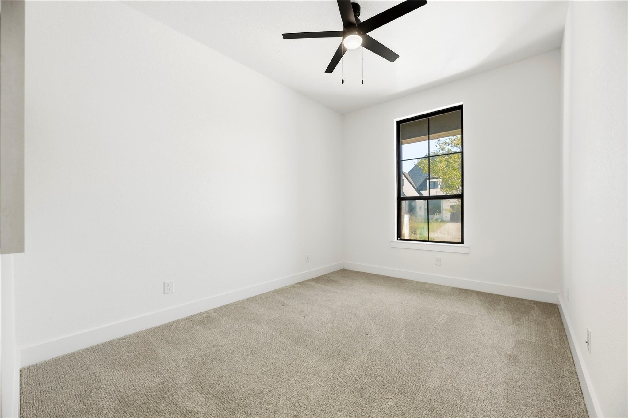 Empty bedroom with beige carpet, a black ceiling fan, and a tall black-framed window.