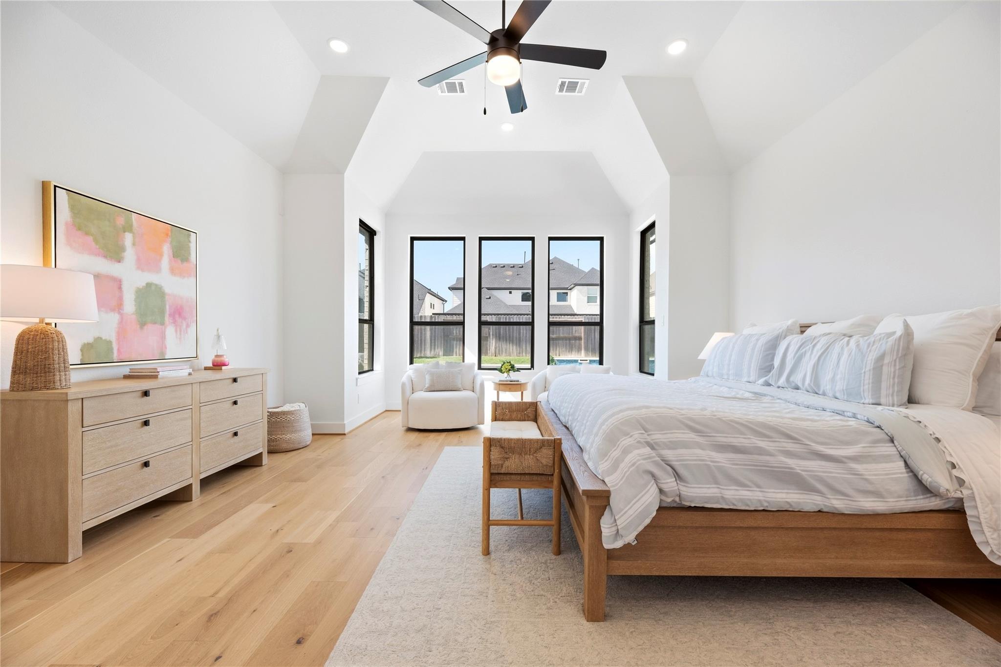 Furnished bedroom with a vaulted ceiling, large windows, striped bedding, and light wood floors.