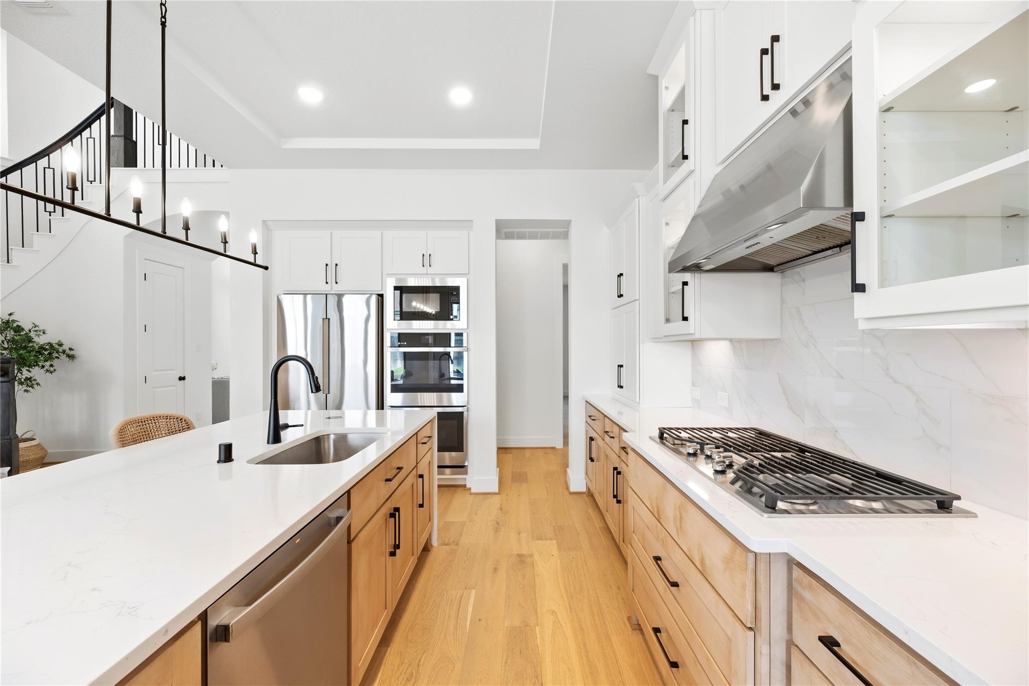 Bright kitchen with a large white island, light wood cabinets, stainless appliances, and a black-railed staircase.
