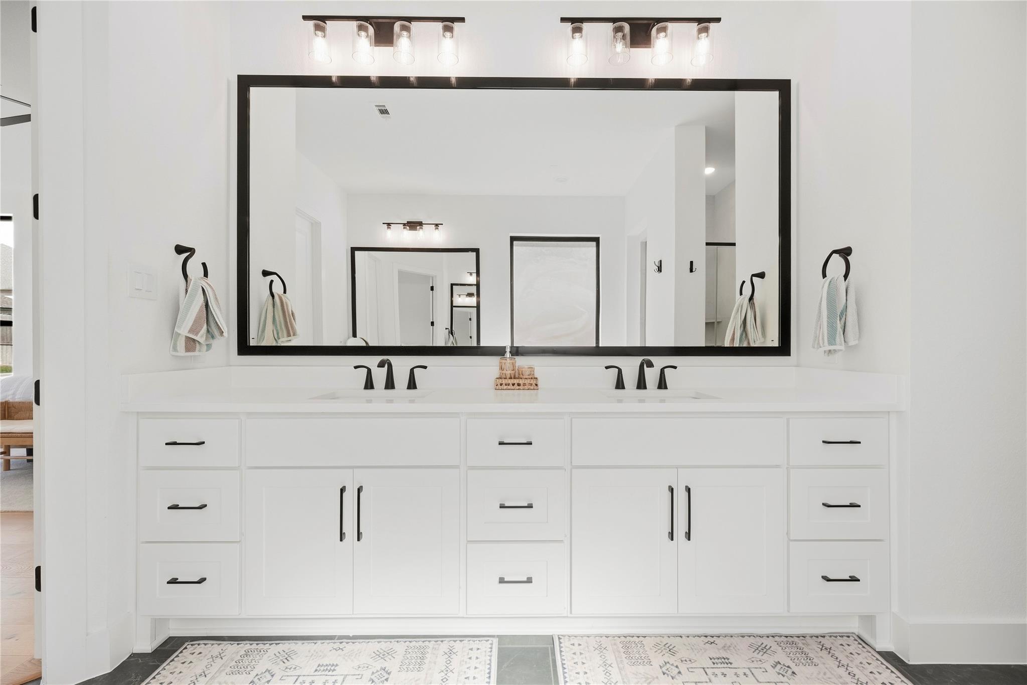 Bathroom with a white double-sink vanity, black-framed mirror, and patterned rugs.