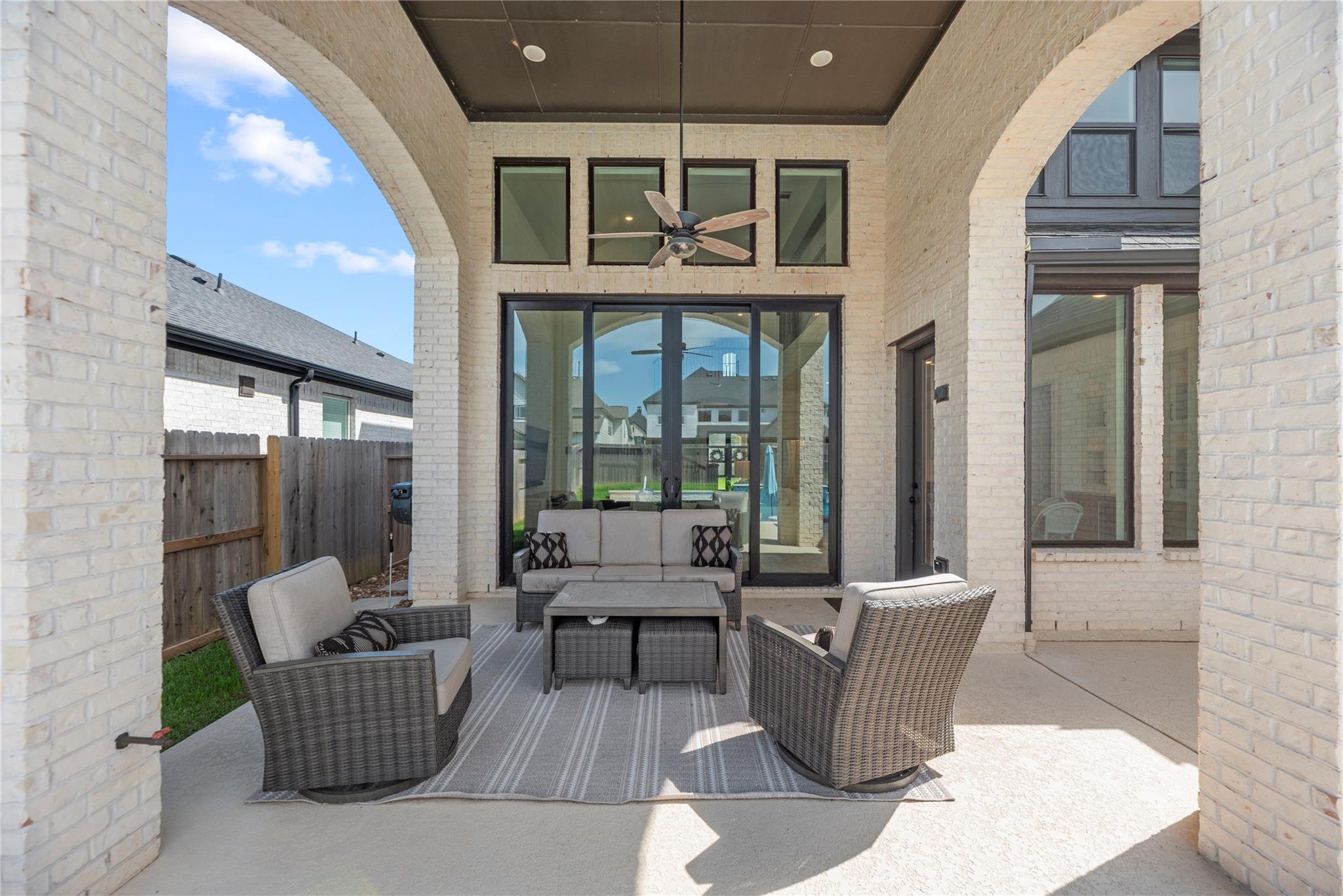 Covered backyard patio with white brick arches, wicker seating, and a ceiling fan.