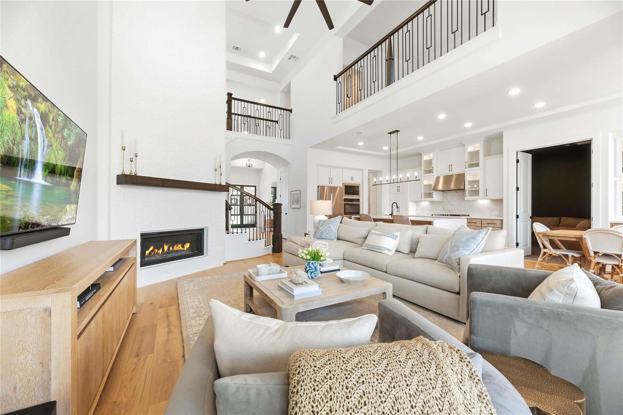 Open living room with white brick fireplace, soaring ceiling, and adjoining kitchen.