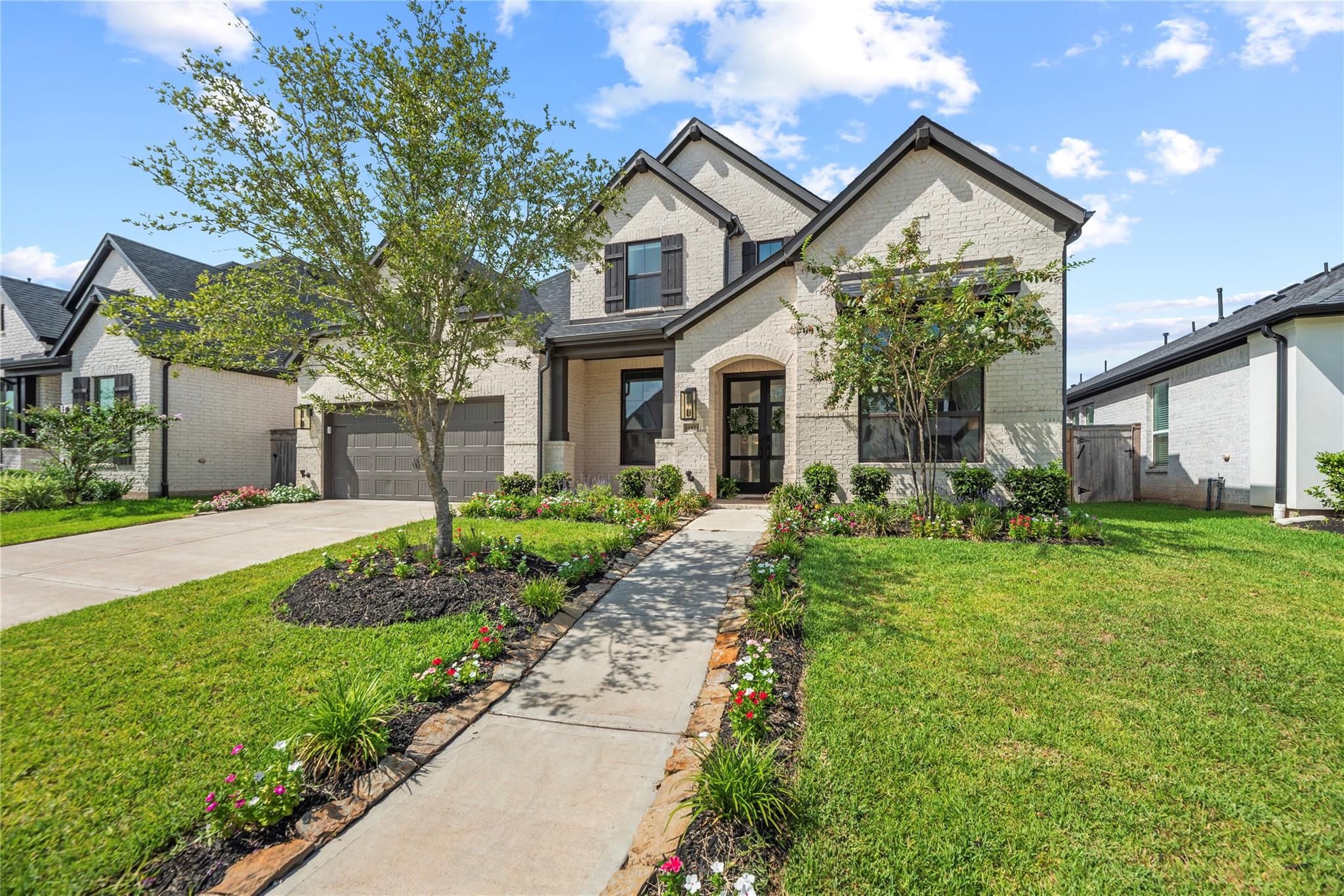 Two-story light brick house with dark trim, a two-car garage, and a flower-lined walkway to the entrance.