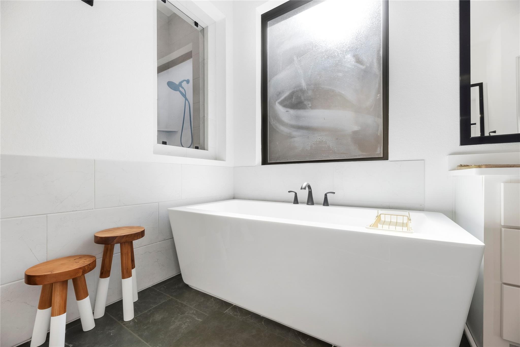 Modern bathroom with a white freestanding soaking tub, dark tile floor, and wooden stools.