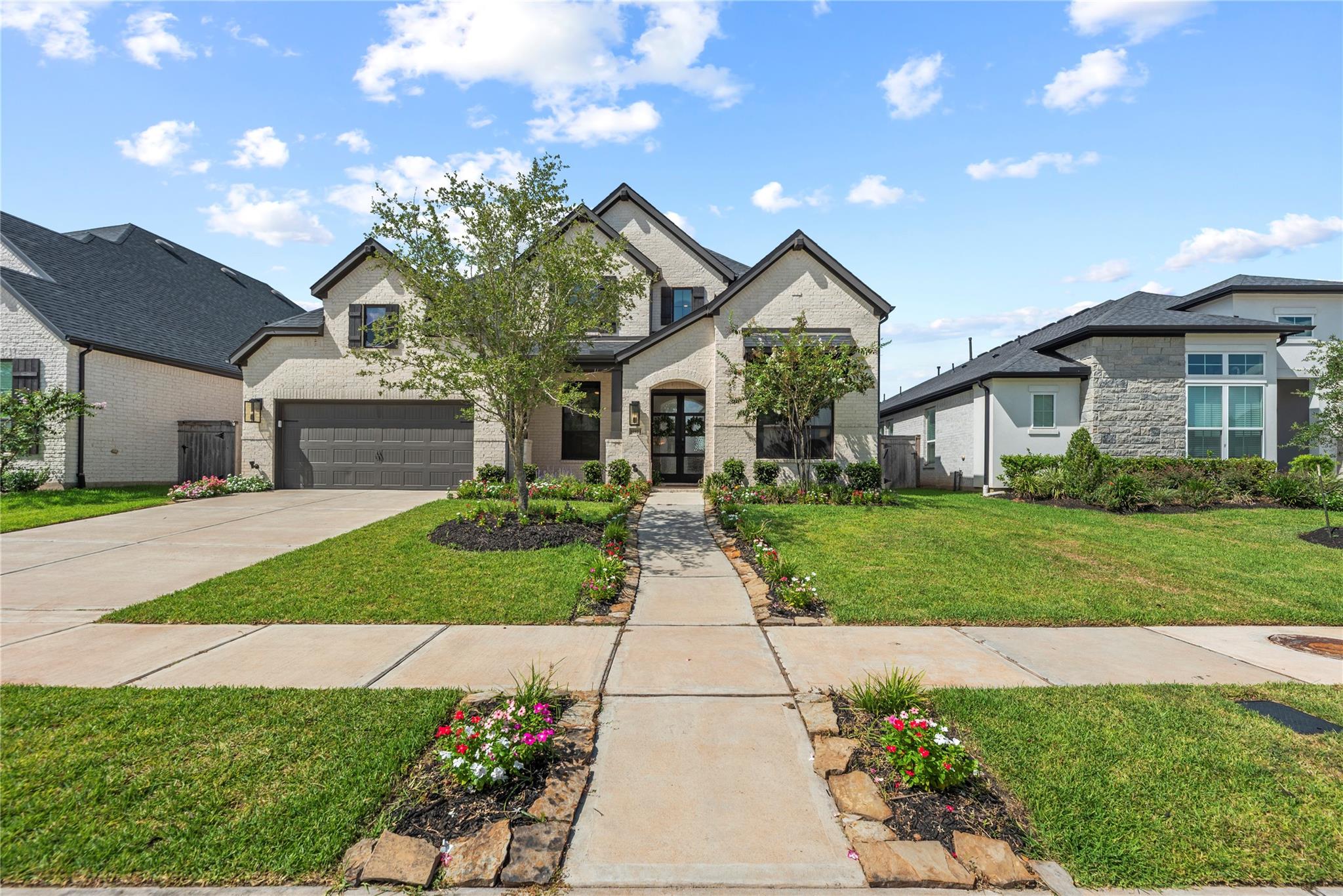 Two-story light brick home with dark roof, double garage, and flower-lined front walkway.