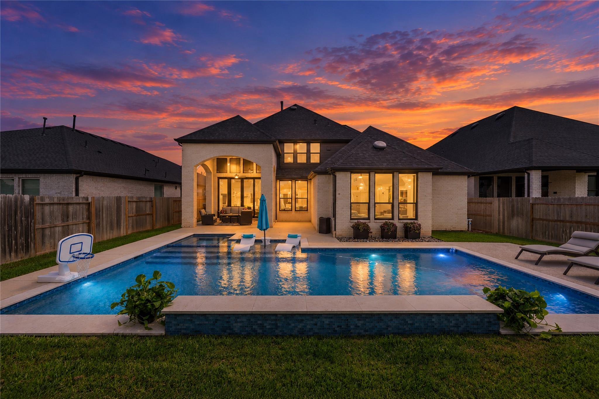 Brick home behind a backyard pool at sunset, with a basketball hoop and lounge chairs.