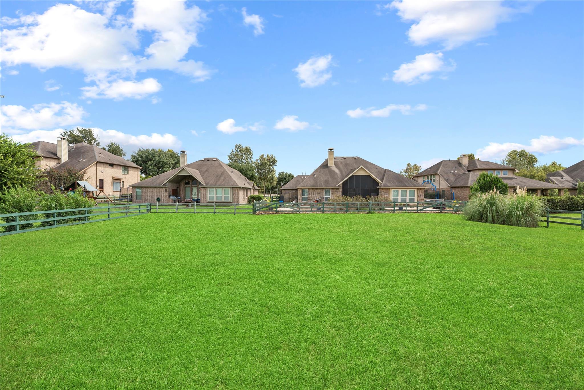 Broad green backyard lawn facing a brick home, with a fenced yard and neighboring houses.