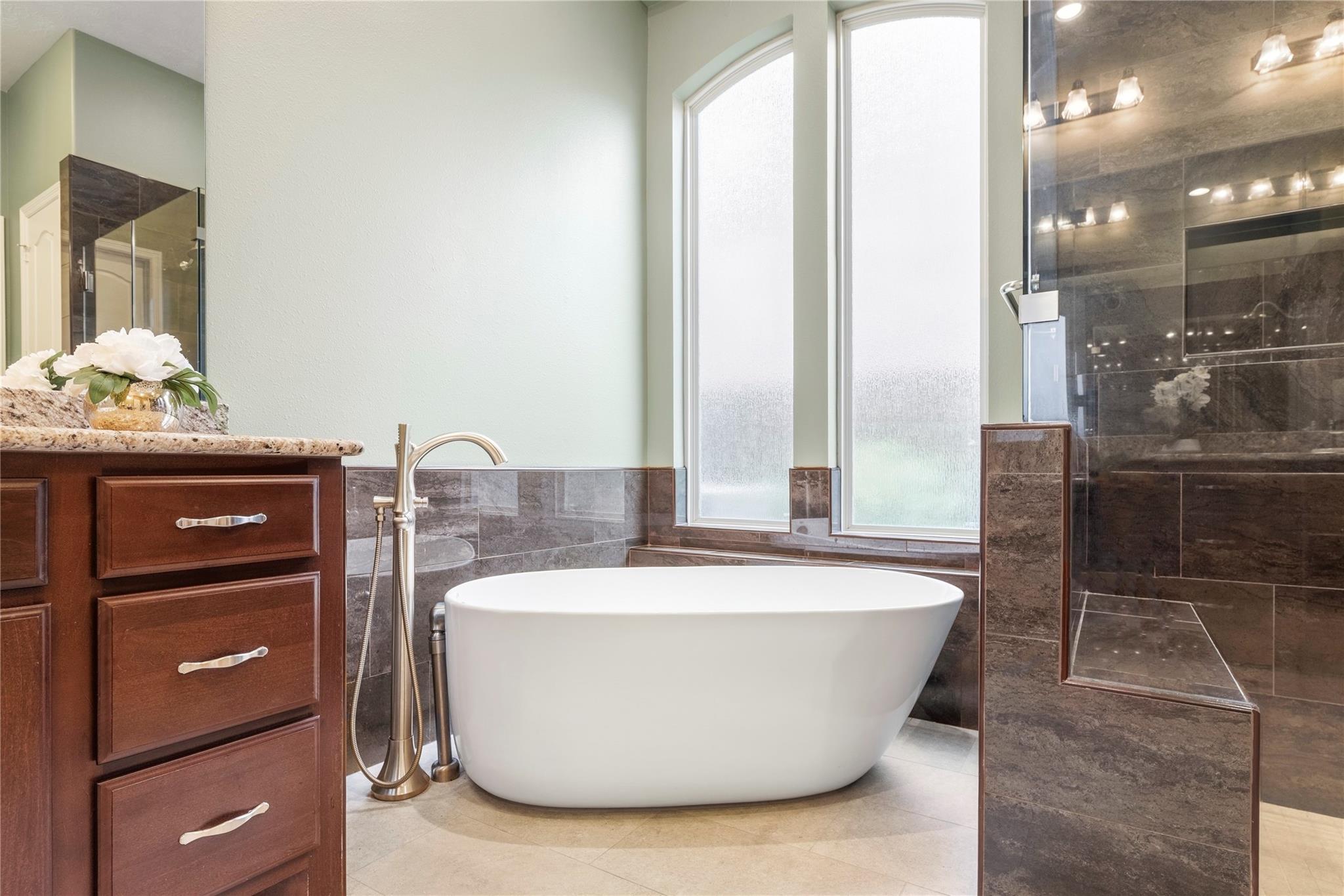 Bathroom with a freestanding soaking tub beneath tall frosted windows beside a dark tiled glass-enclosed shower.