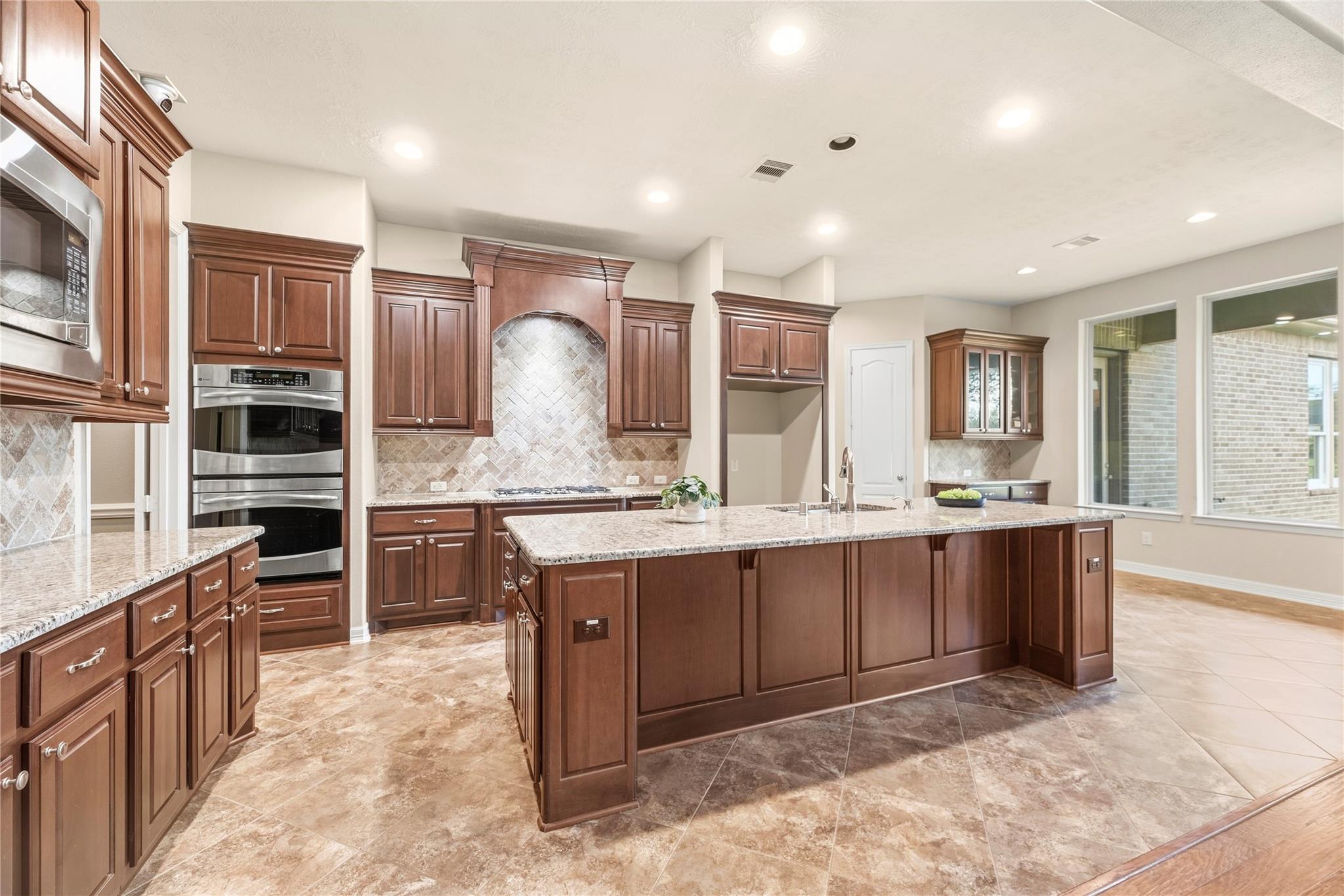 Kitchen with a large granite island, dark wood cabinets, double ovens, and a decorative tile backsplash.