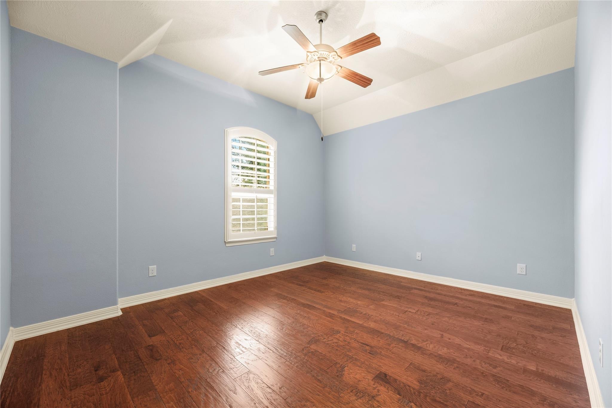 Empty bedroom with dark hardwood floors, vaulted ceiling, ceiling fan, and arched shuttered window.