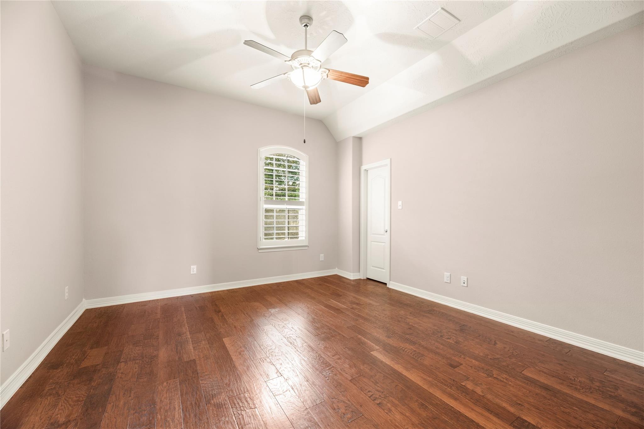 Empty bedroom with hardwood floors, vaulted ceiling, ceiling fan, and arched shuttered window.