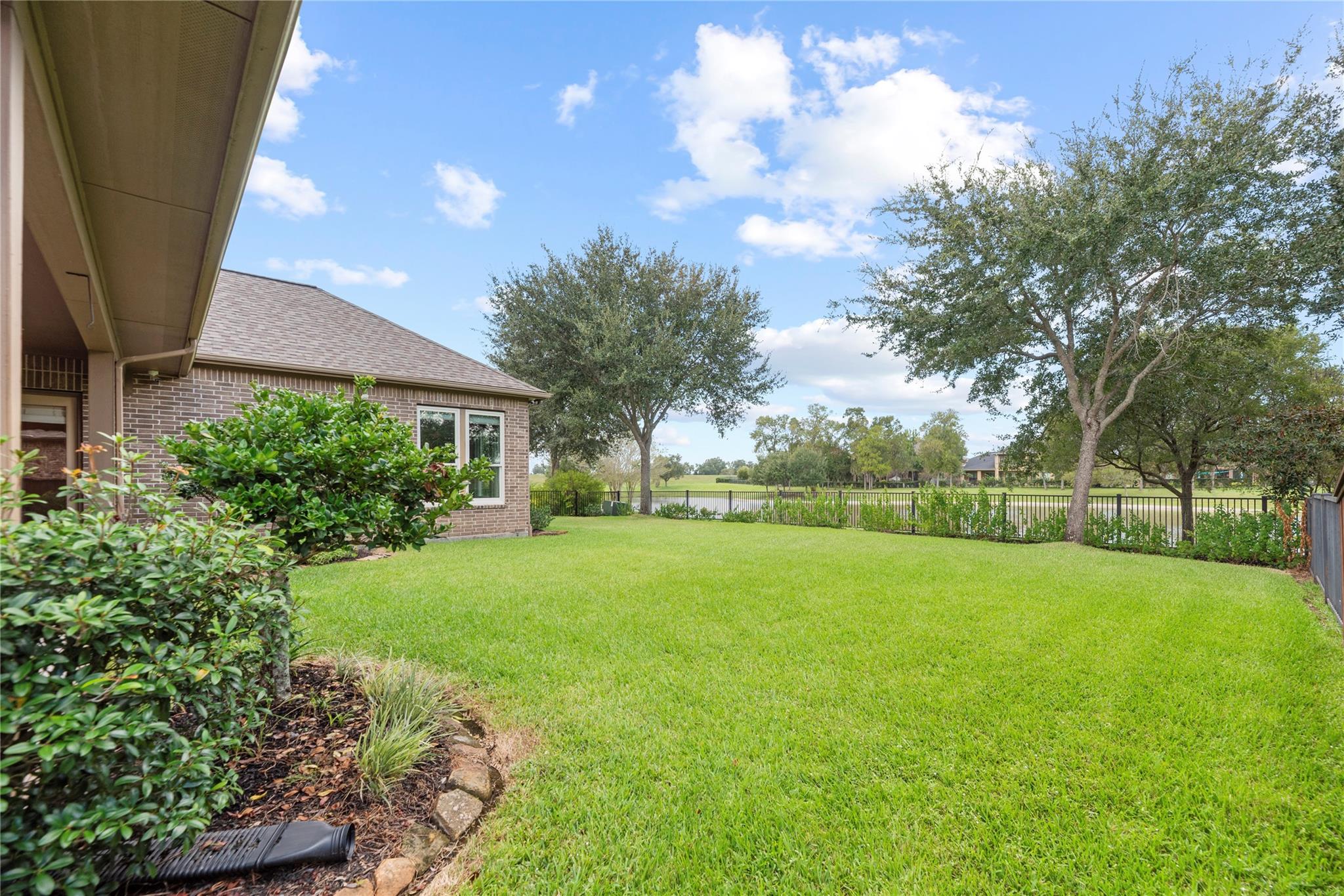 Brick home’s spacious backyard with a broad lawn, mature trees, and a fenced open-space view.
