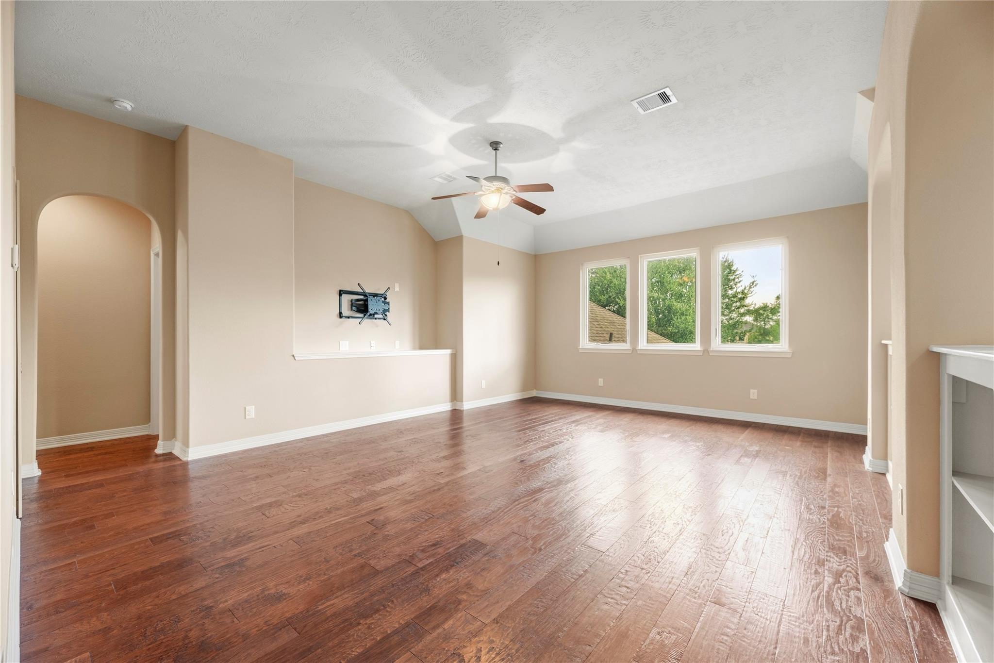 Empty living room with hardwood floors, vaulted ceiling, ceiling fan, and three windows.
