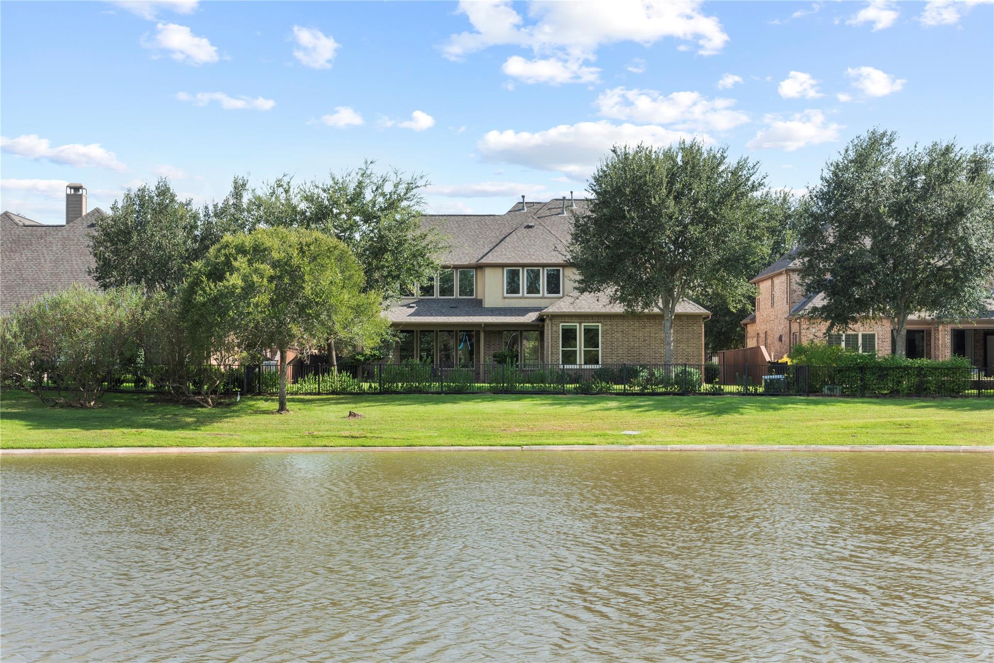 Two-story home across a fenced lawn from a lake, shaded by mature trees.