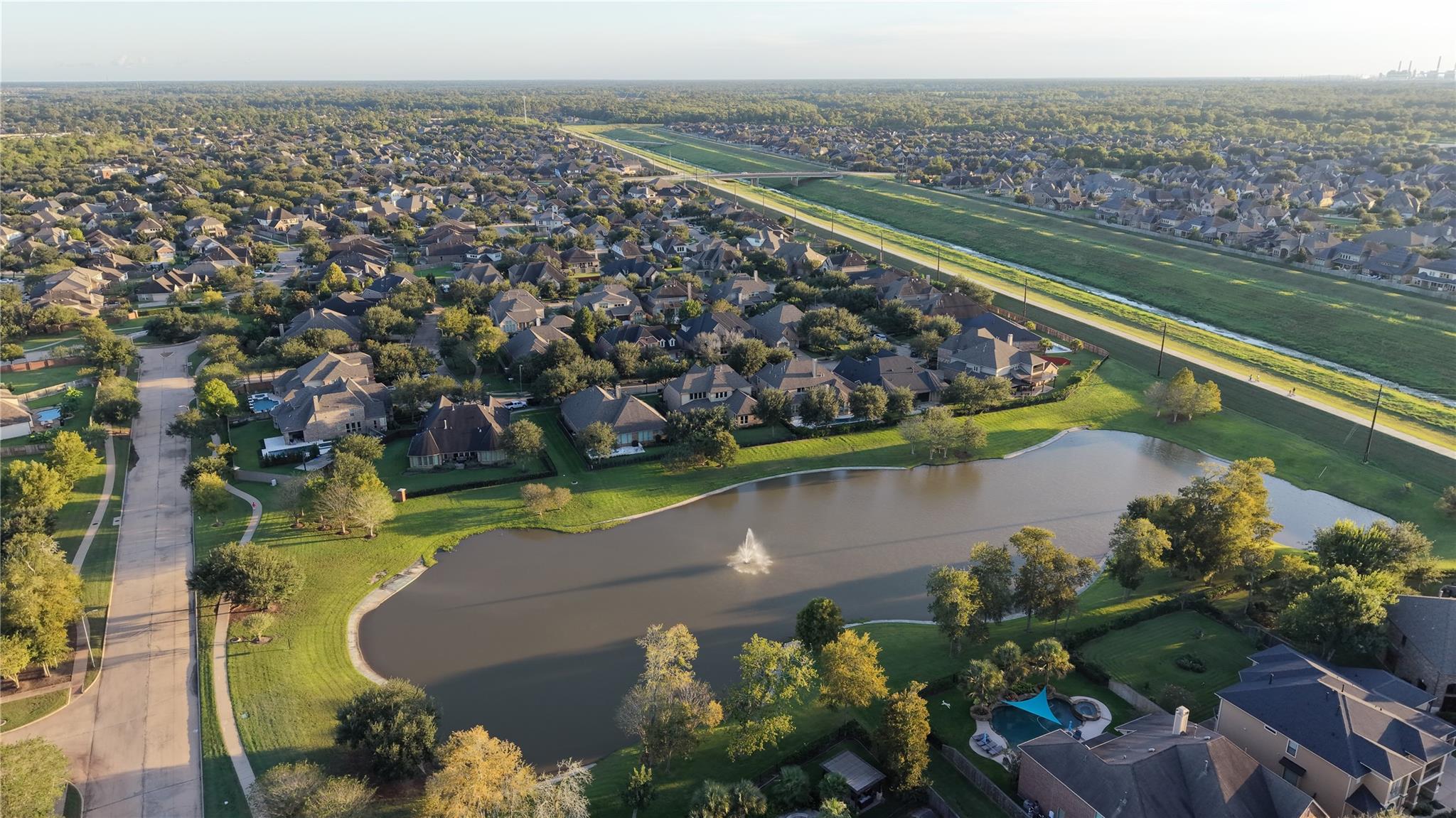 Aerial view of homes around a pond with a fountain and a long grassy drainage channel.