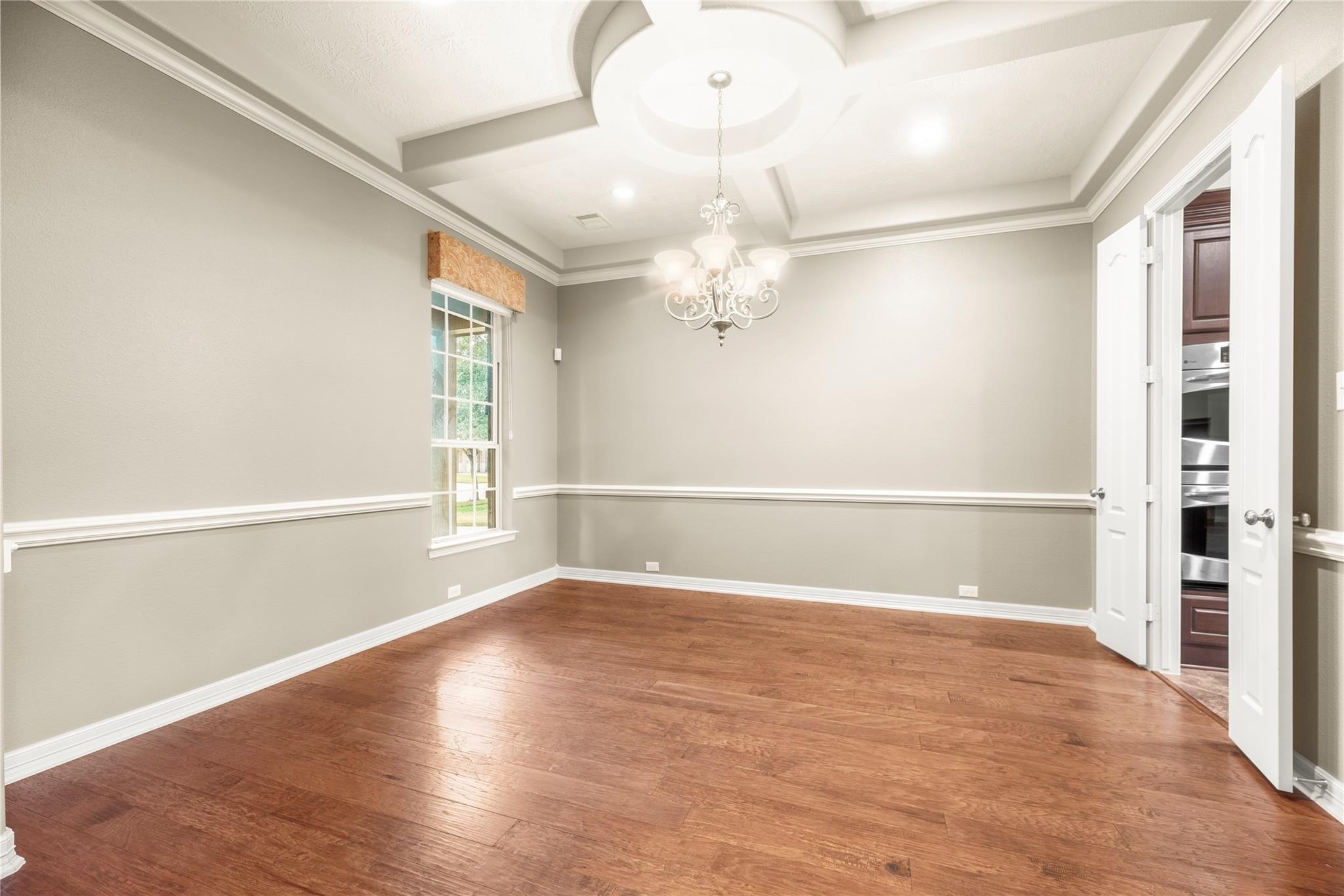 Empty dining room with hardwood floors, a tray ceiling, chandelier, and open kitchen doors.