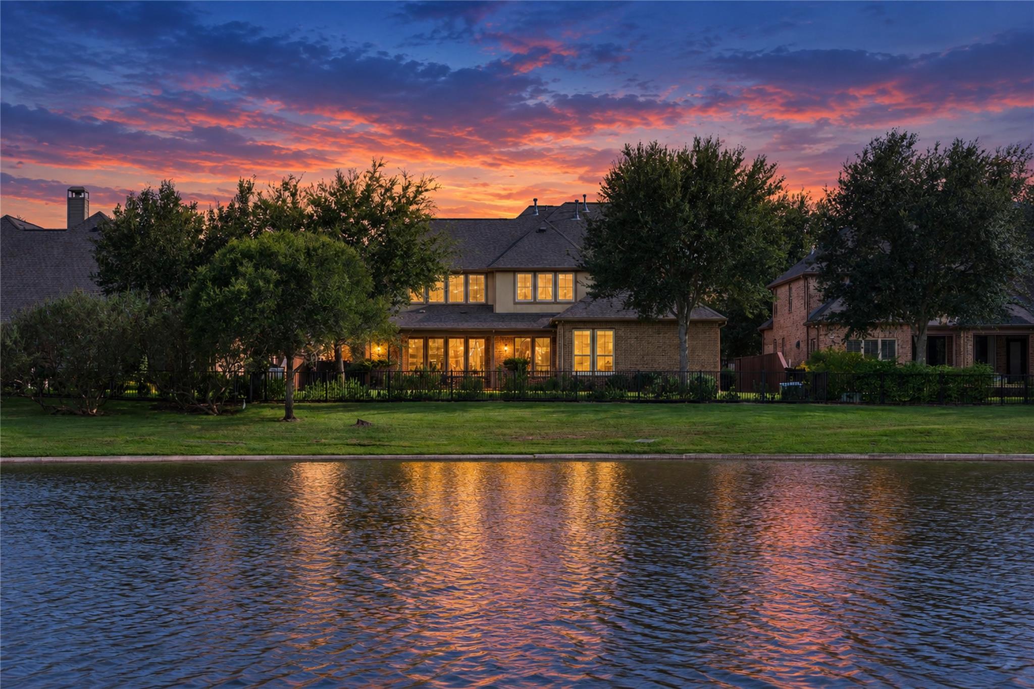 Two-story brick home with glowing windows across the water beneath a colorful dusk sky.