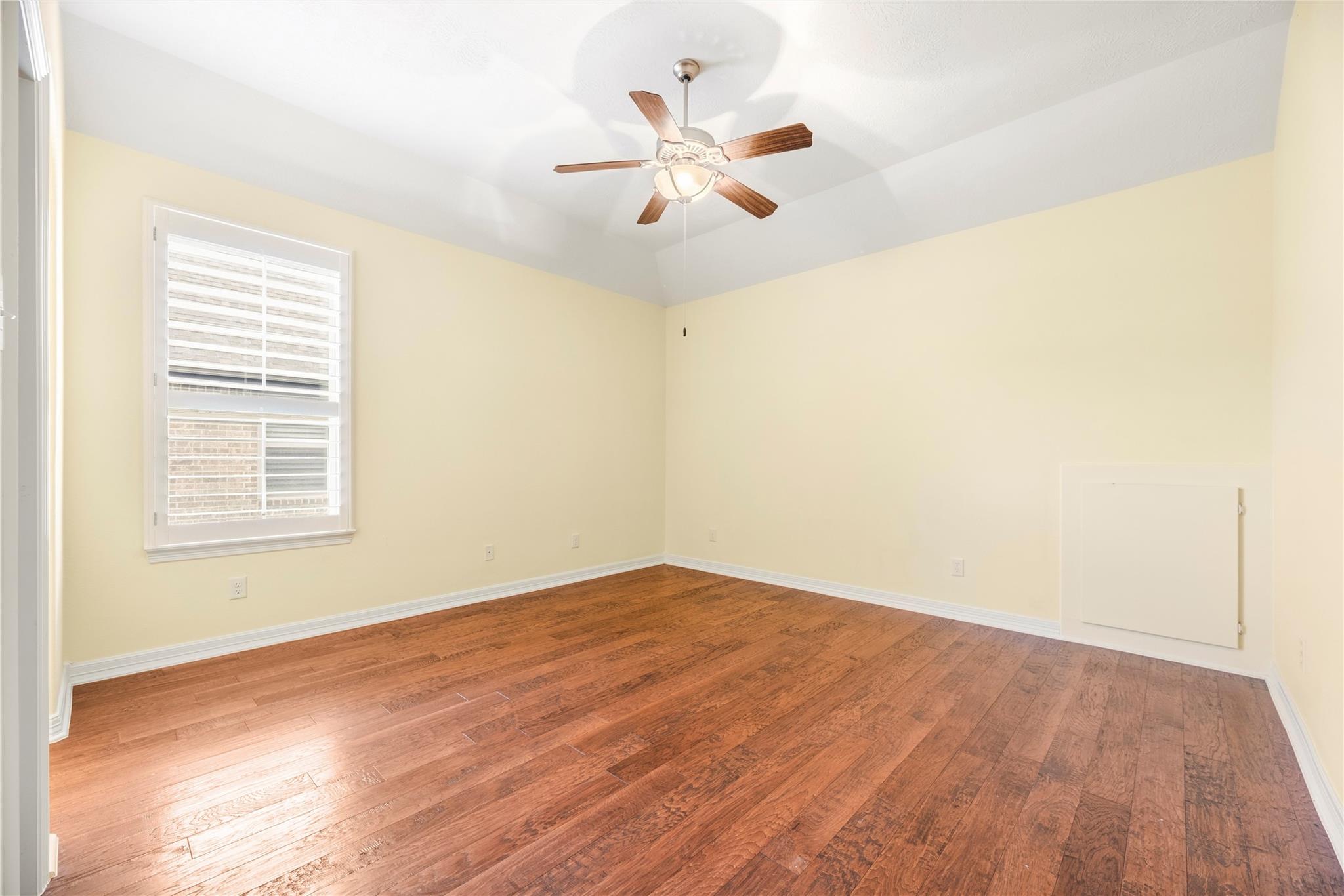 Empty bedroom with hardwood floors, shuttered window, and wooden-blade ceiling fan.