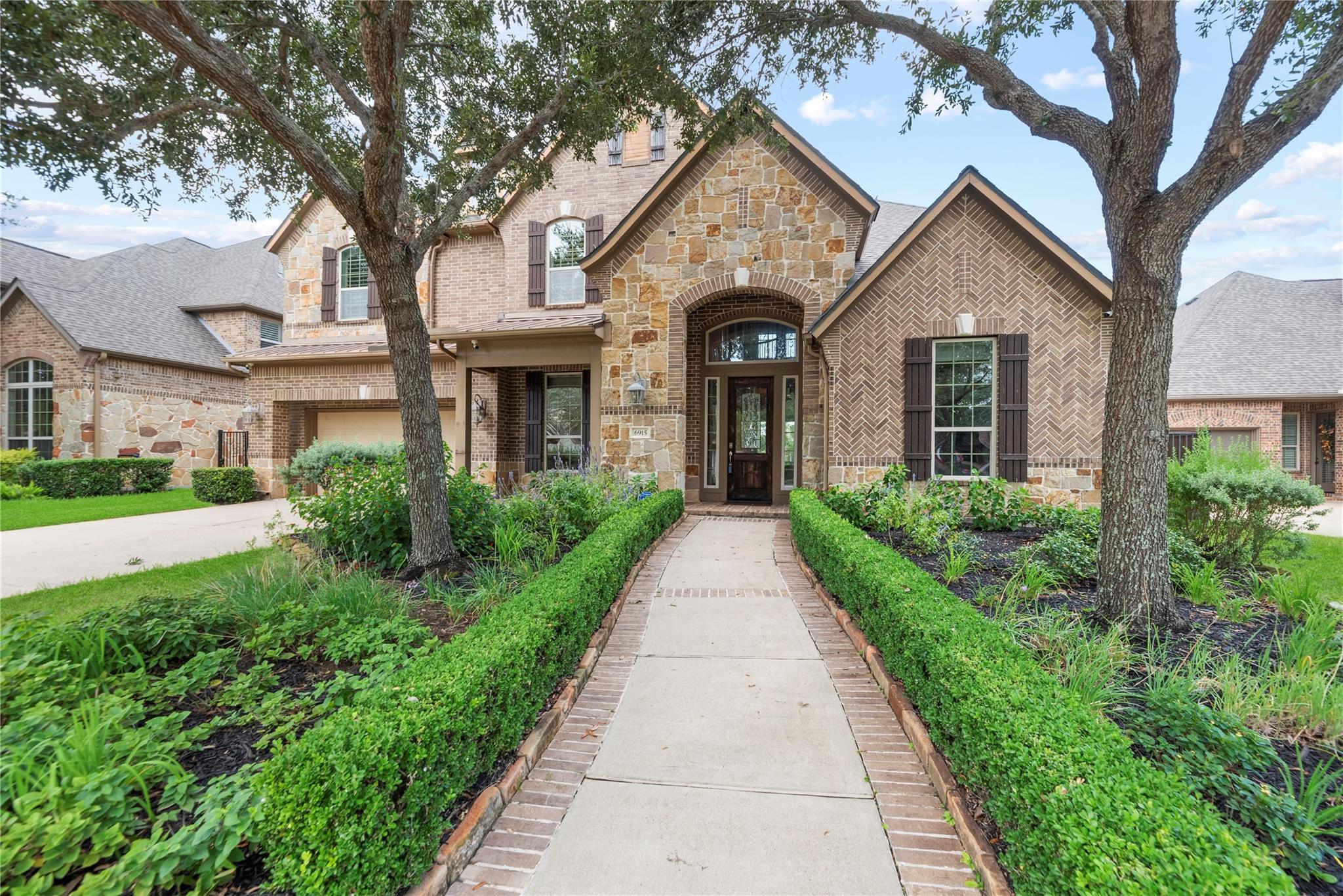 Two-story stone-and-brick home with an arched entry, mature trees, and a landscaped walkway.
