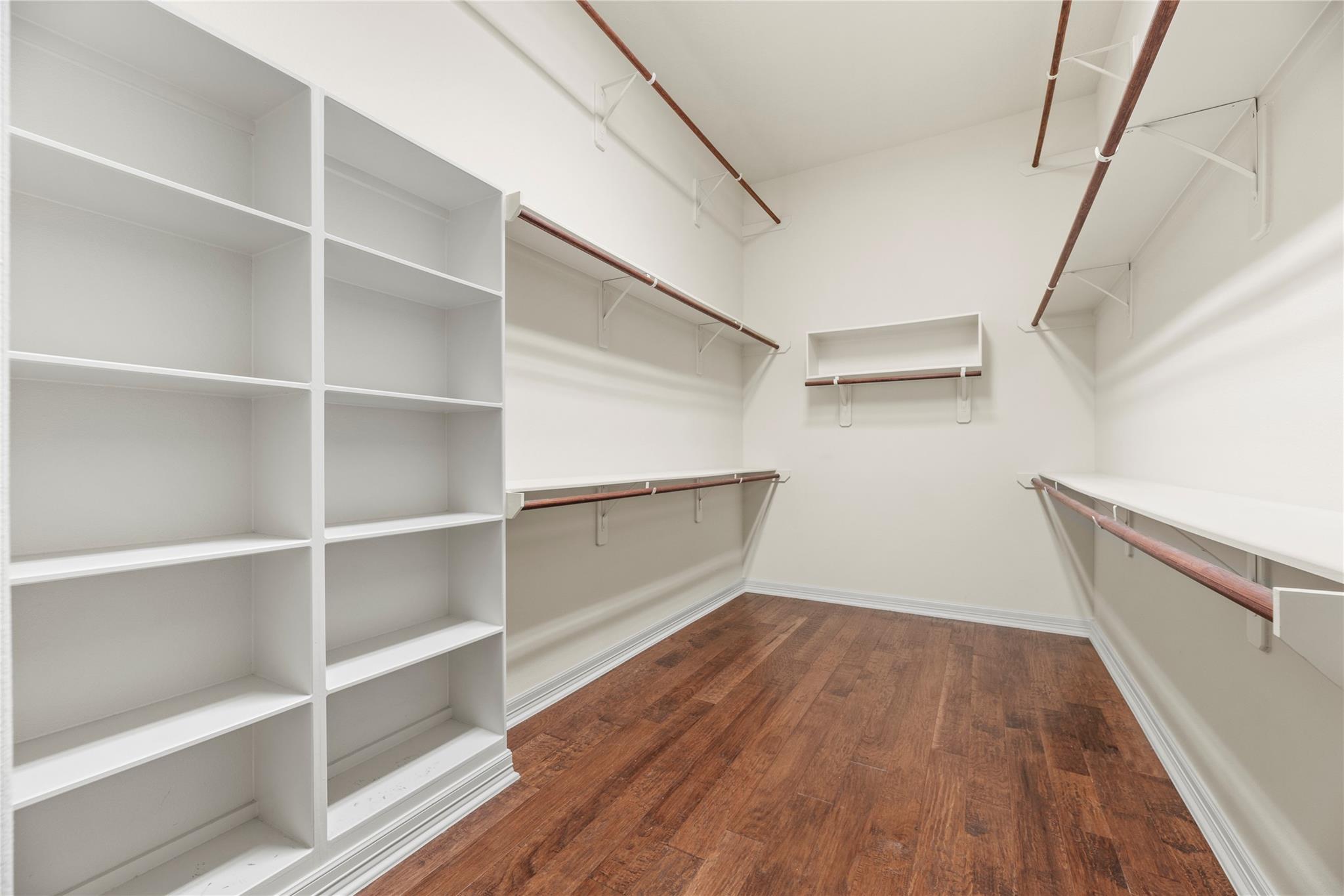 Empty walk-in closet with white shelving, long hanging rods, and warm wood flooring.