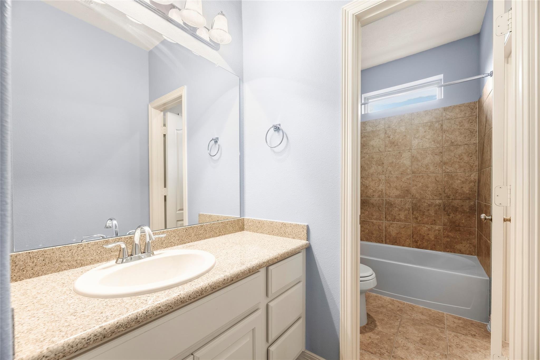 Bathroom with a granite vanity and mirror beside a tub with a brown tiled surround.