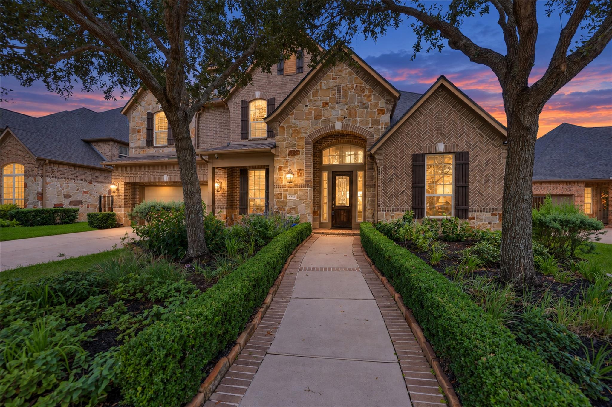 Two-story brick and stone home at dusk, framed by mature trees and hedges along the front walkway.