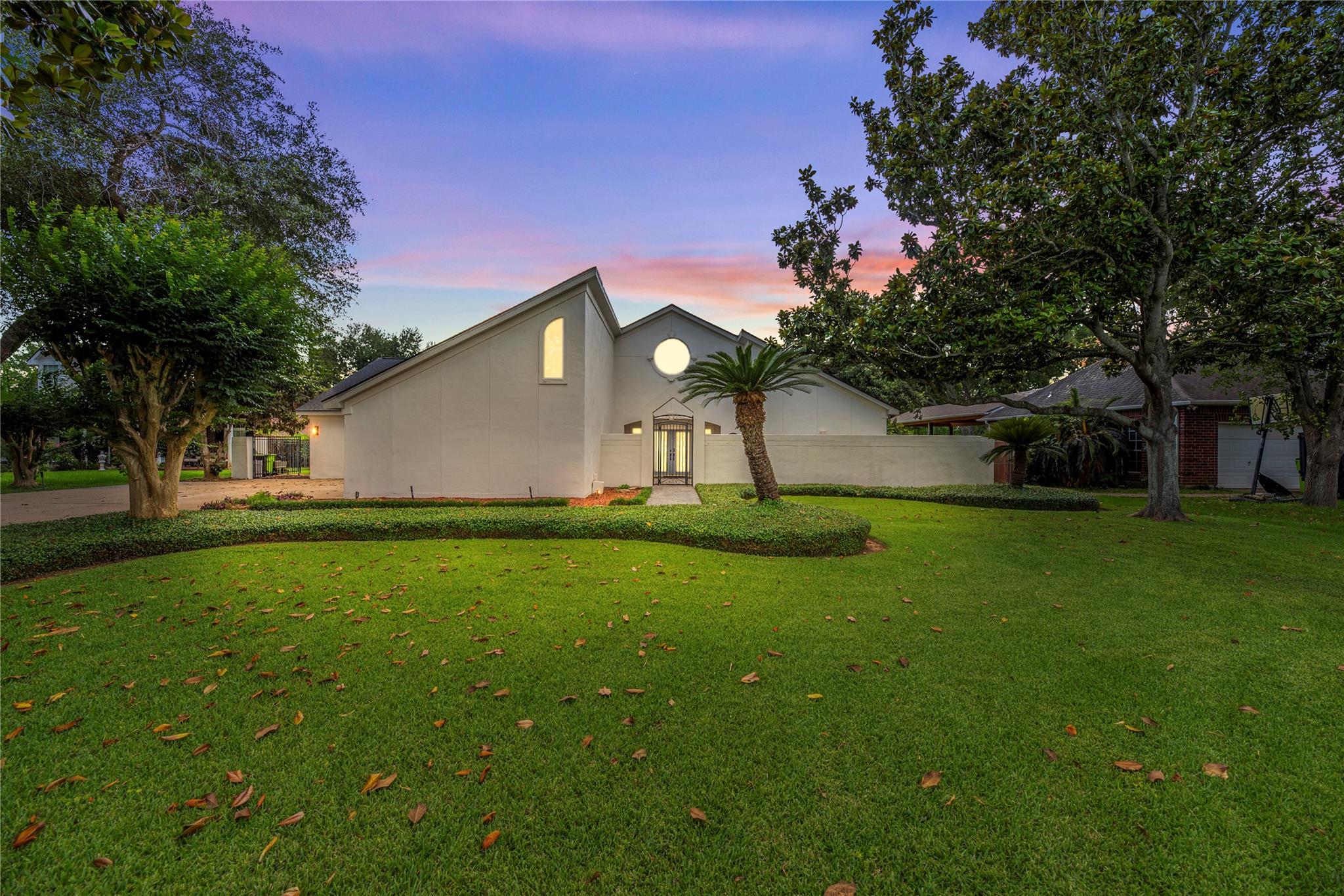 Light-colored home and gated entrance behind a manicured lawn, framed by mature trees at dusk.