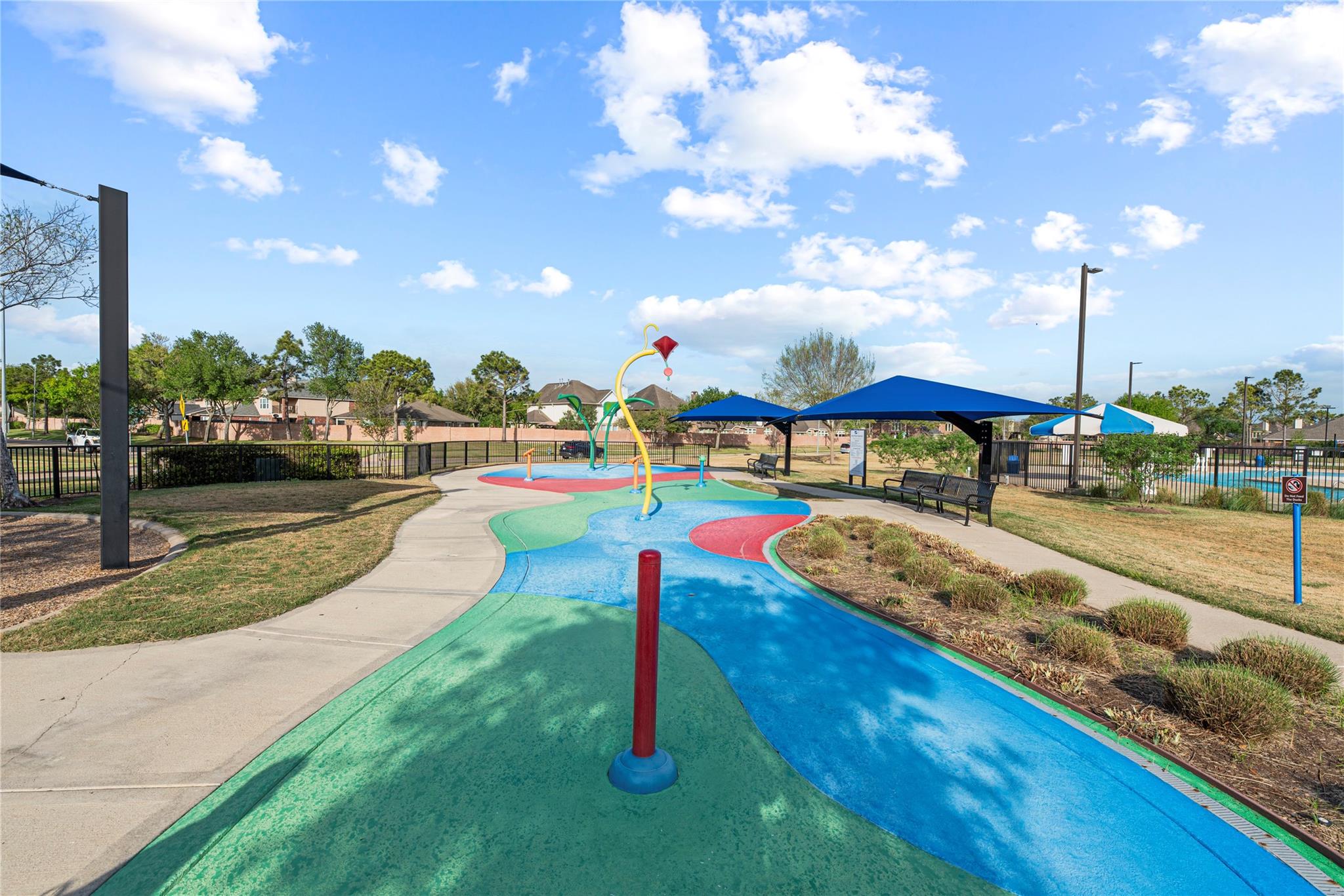 Community splash pad with colorful water features, shade canopies, benches, and a fenced pool nearby.