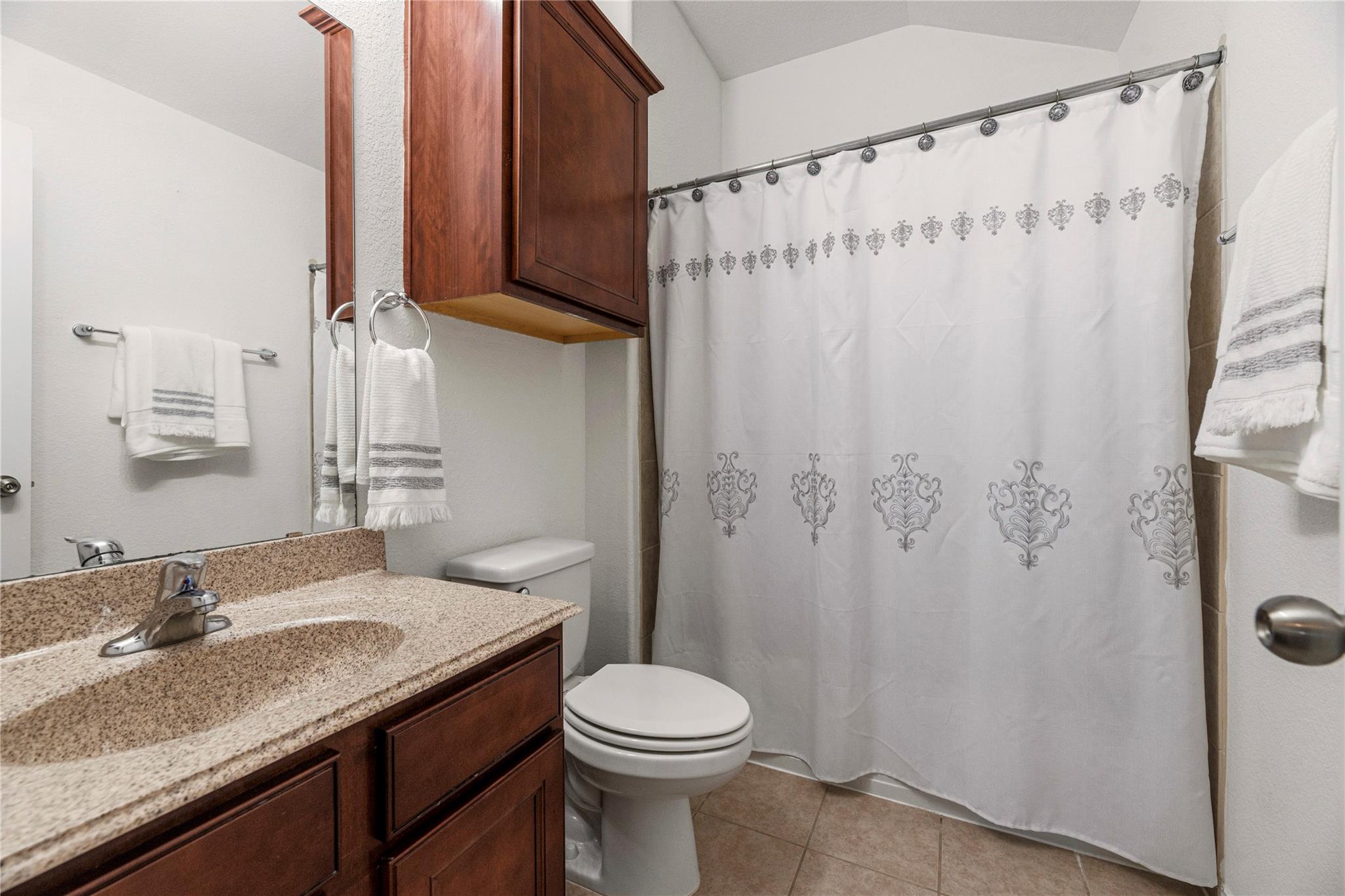 Bathroom with a granite vanity, dark wood cabinets, toilet, and patterned white shower curtain.