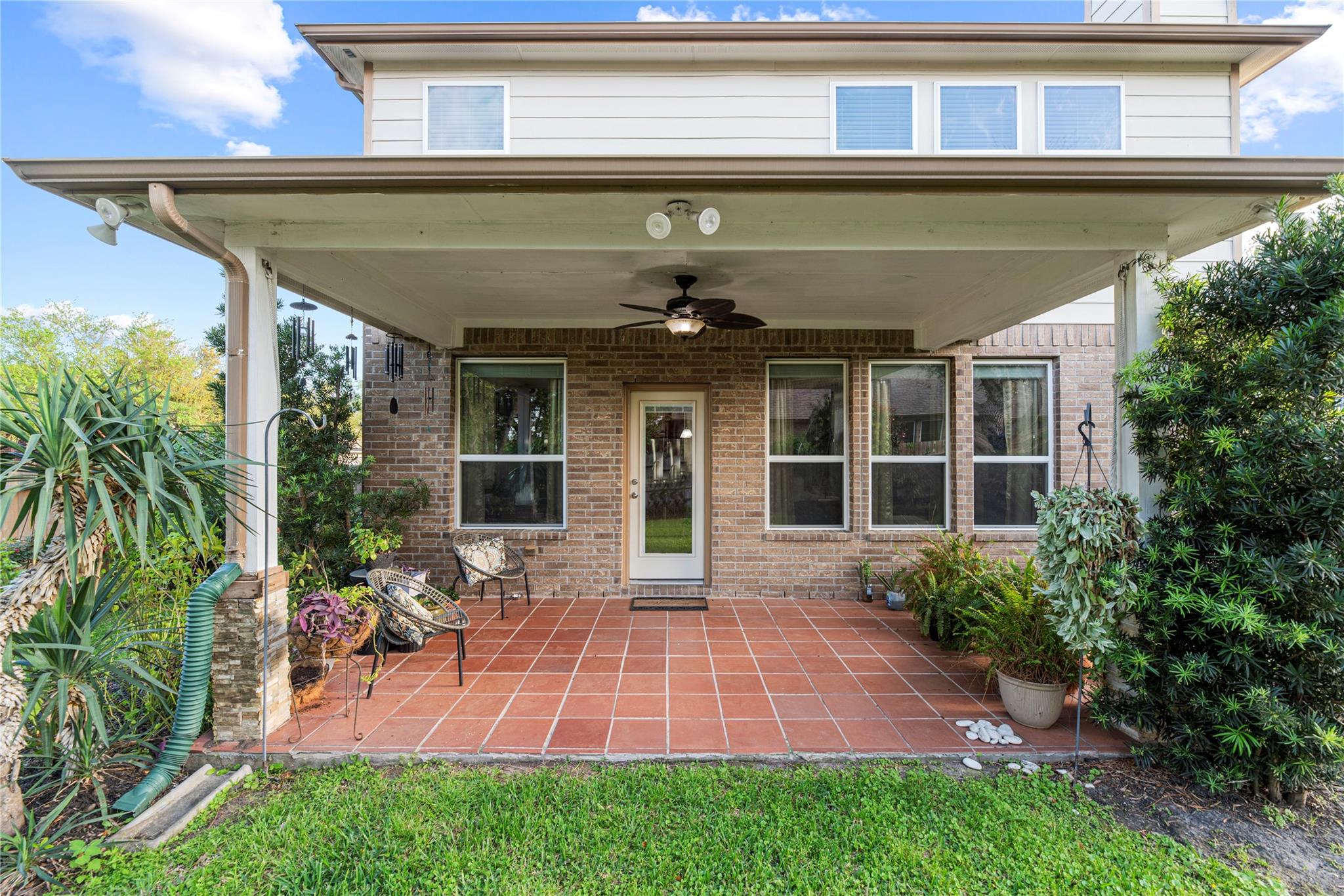 Covered brick-home patio with terracotta tiles, outdoor chairs, ceiling fan, and lush garden plants.