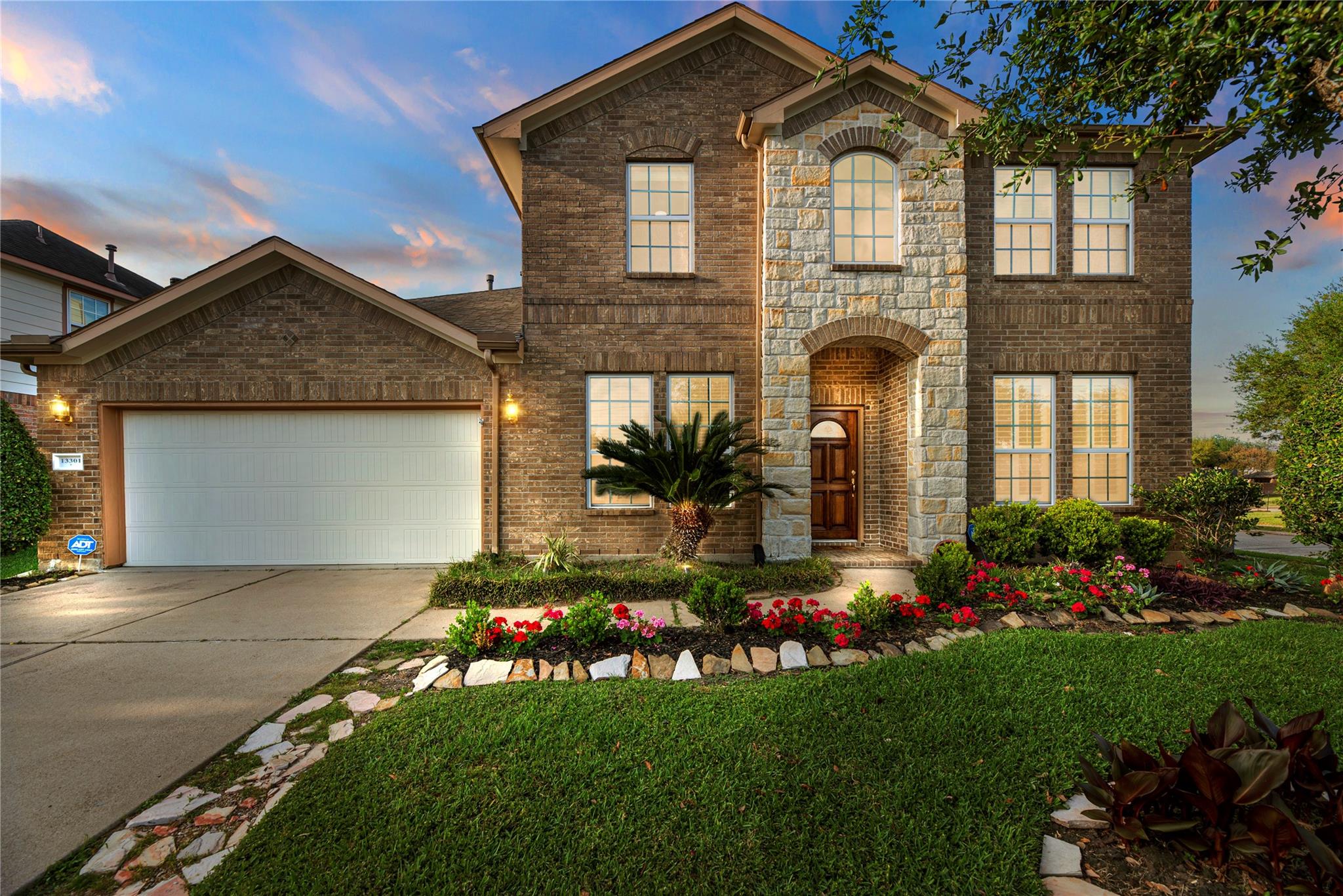Two-story brick home with a stone entry, two-car garage, and colorful landscaped flower beds.