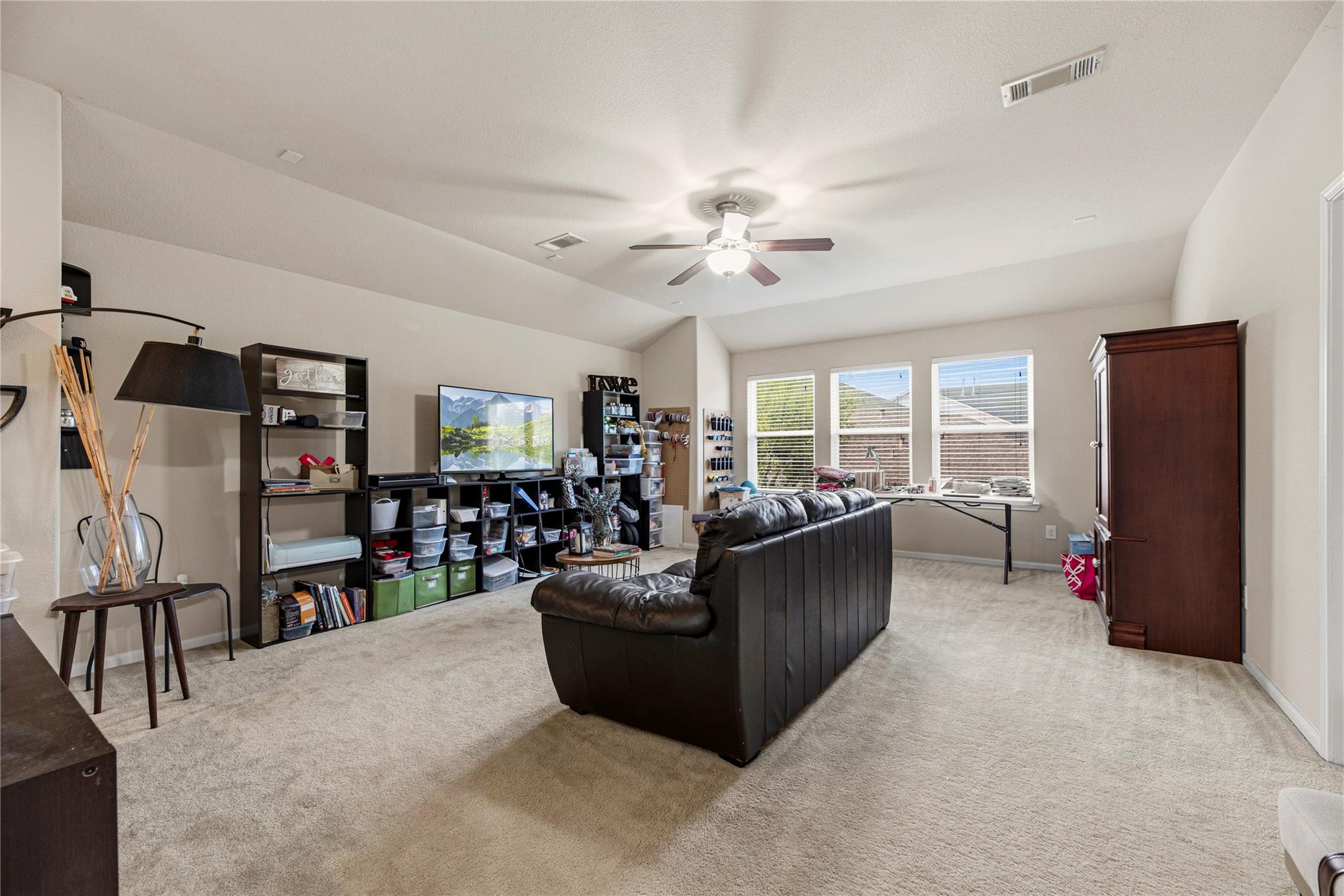 Furnished living room with a dark leather sectional, ceiling fan, and three bright windows.