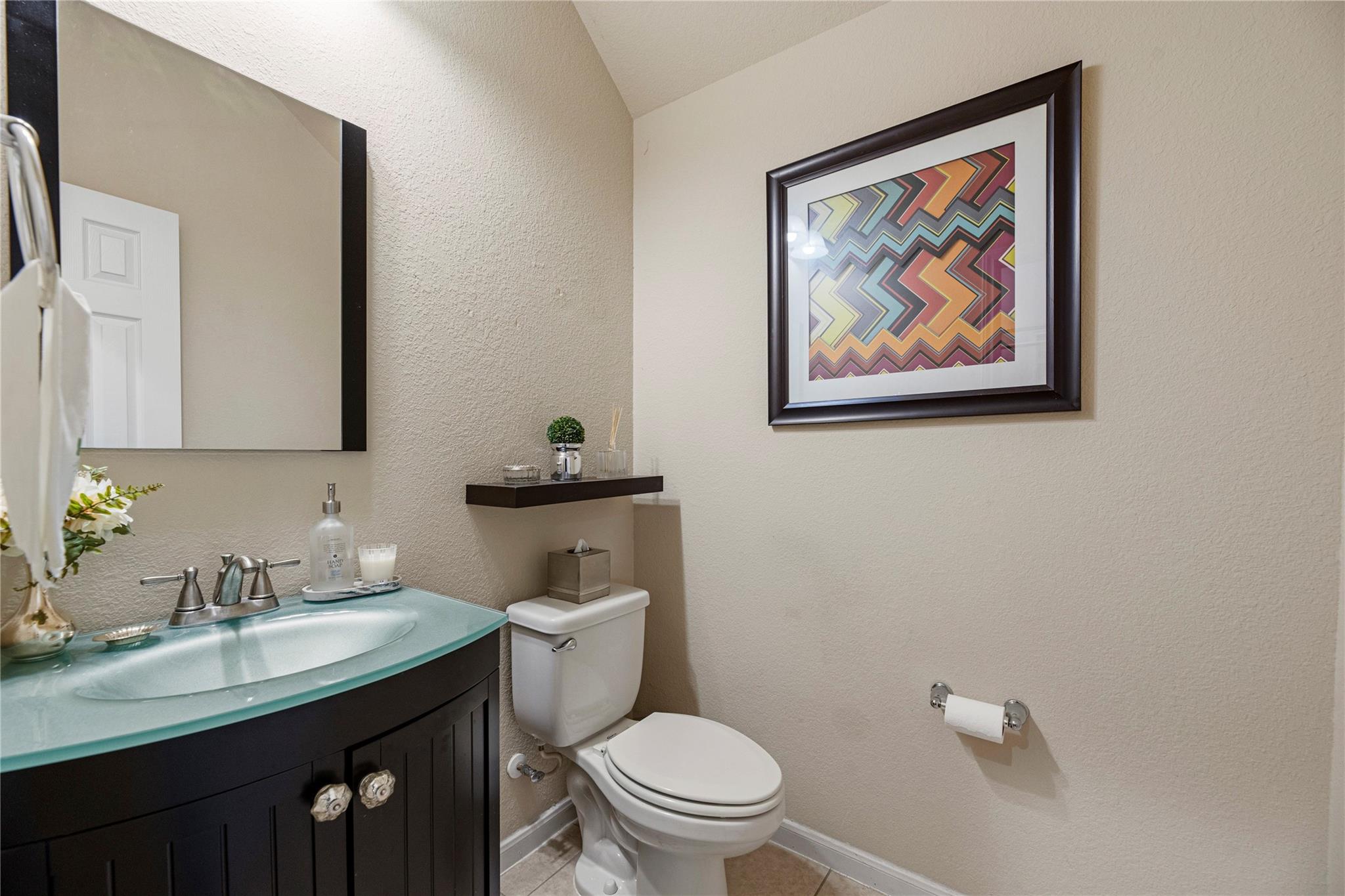 Bathroom with a turquoise glass vessel sink, dark vanity, and colorful framed artwork above the toilet.