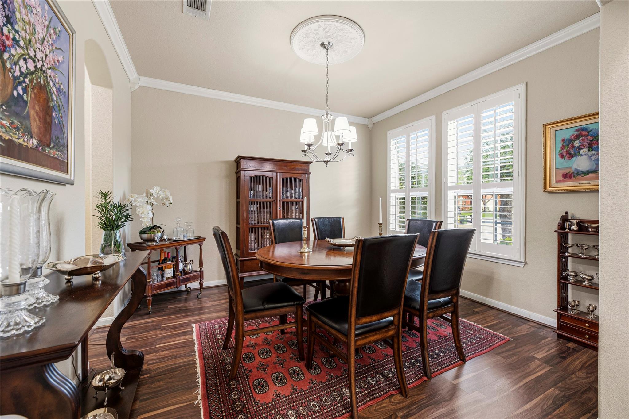 Dining room with a round wood table, plantation-shuttered windows, and a red patterned rug.