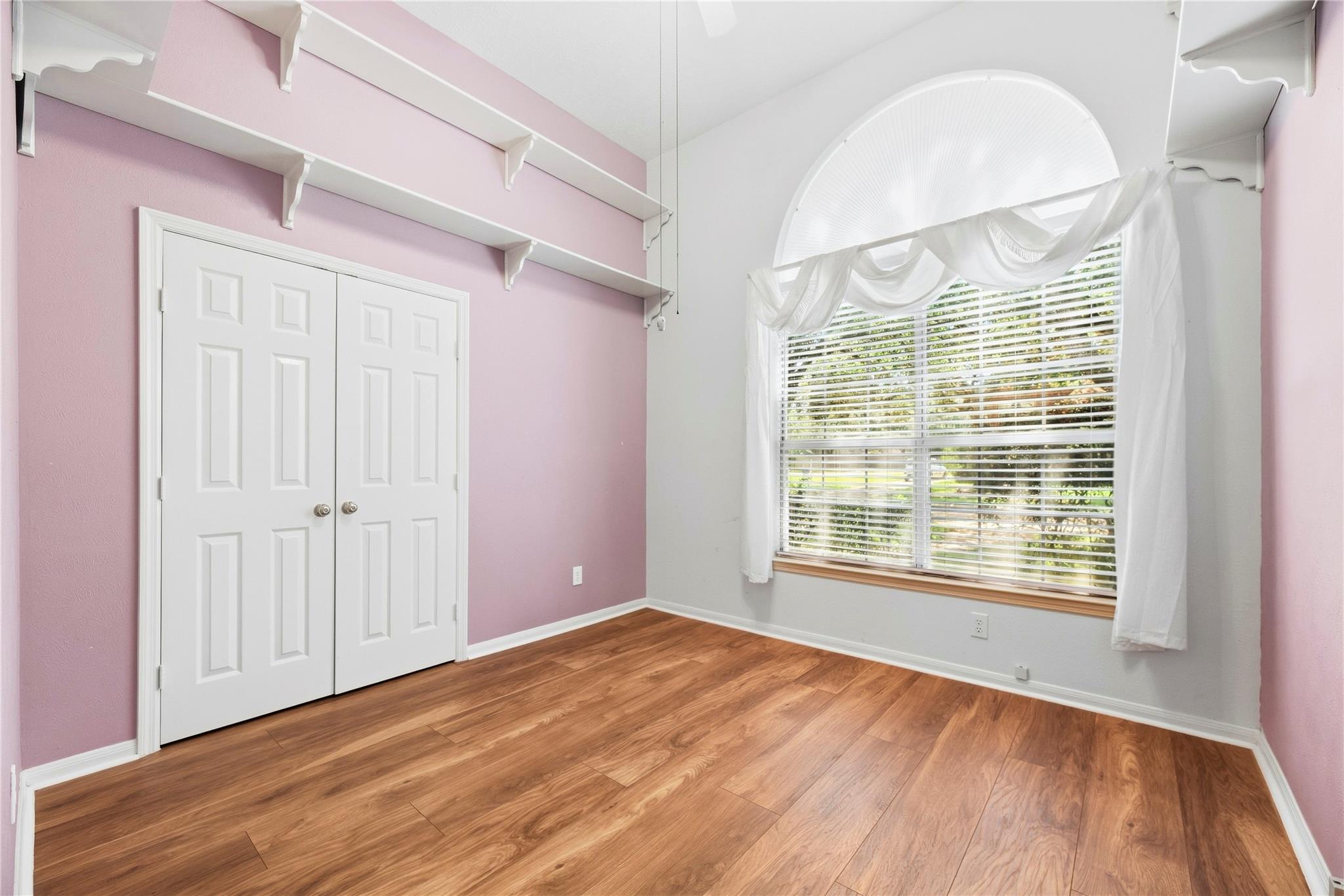 Empty bedroom with pink walls, wood-look flooring, and a large arched window.