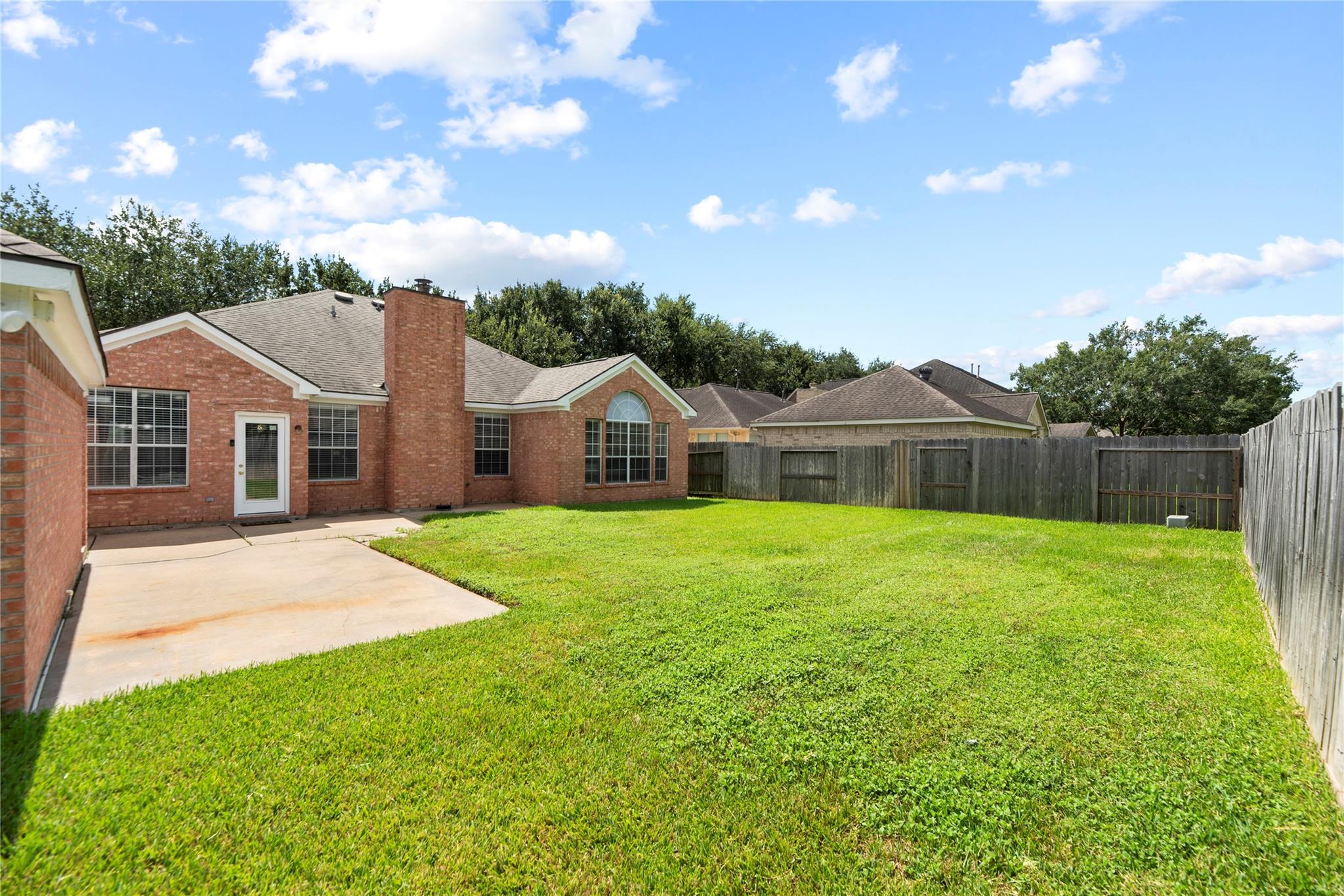 Backyard with a broad lawn, concrete patio, red-brick house, and wooden privacy fence.