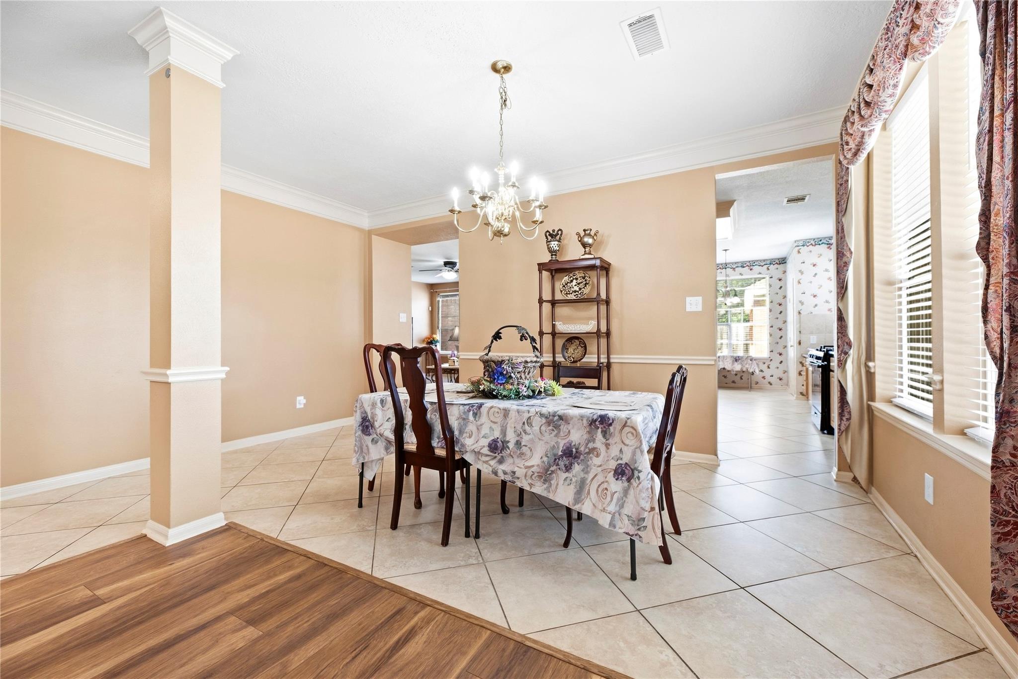 Dining room with a floral-covered table, chandelier, tiled floor, and large curtained windows.