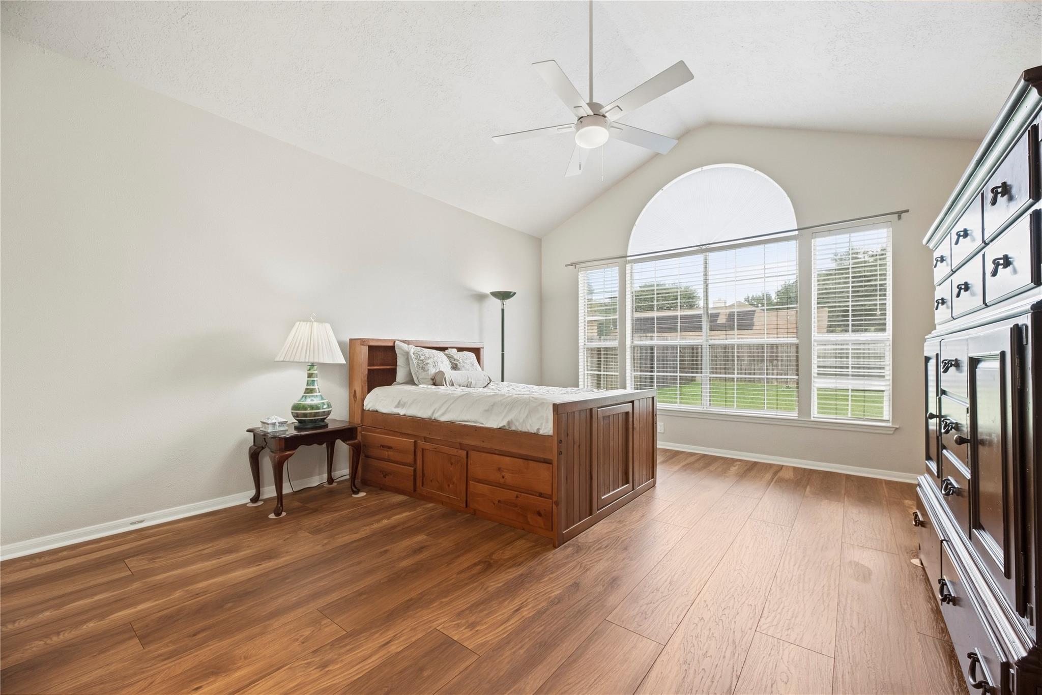 Bedroom with a wooden storage bed, warm wood flooring, and a large arched window.