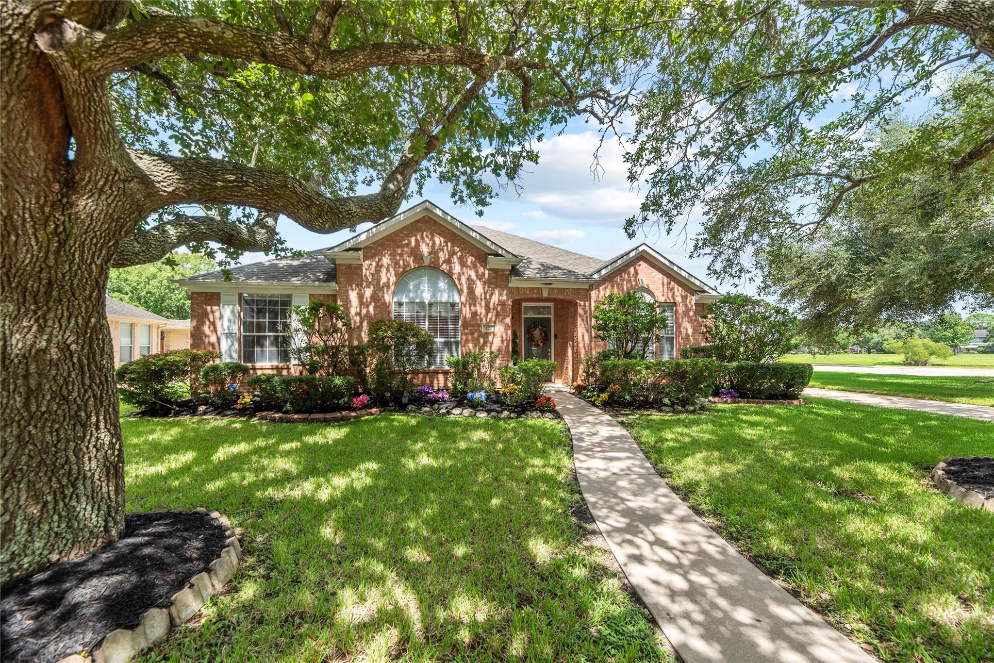 Red-brick house with arched windows, landscaped flower beds, and a walkway beneath a large oak tree.